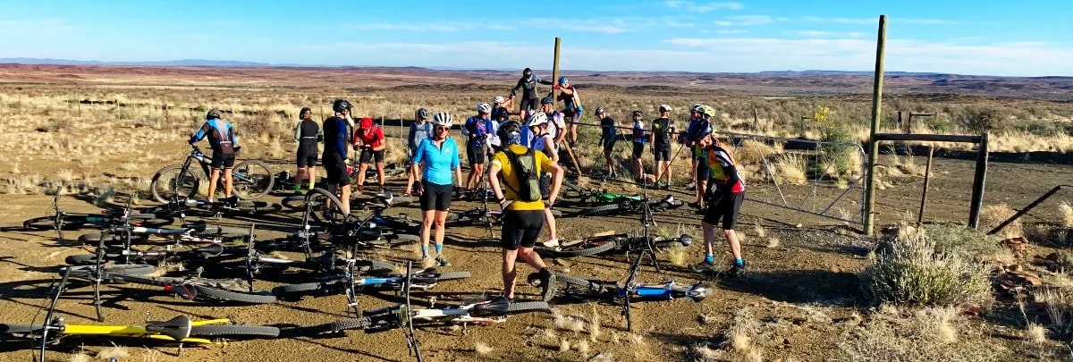 Group of cyclists with bicycles resting and preparing at a desert trail in a wide open landscape under a clear blue sky.