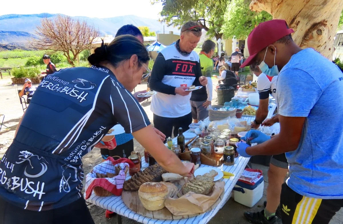 People at an outdoor food stall with bread, jars, and condiments, under a tree, with mountains in the background.