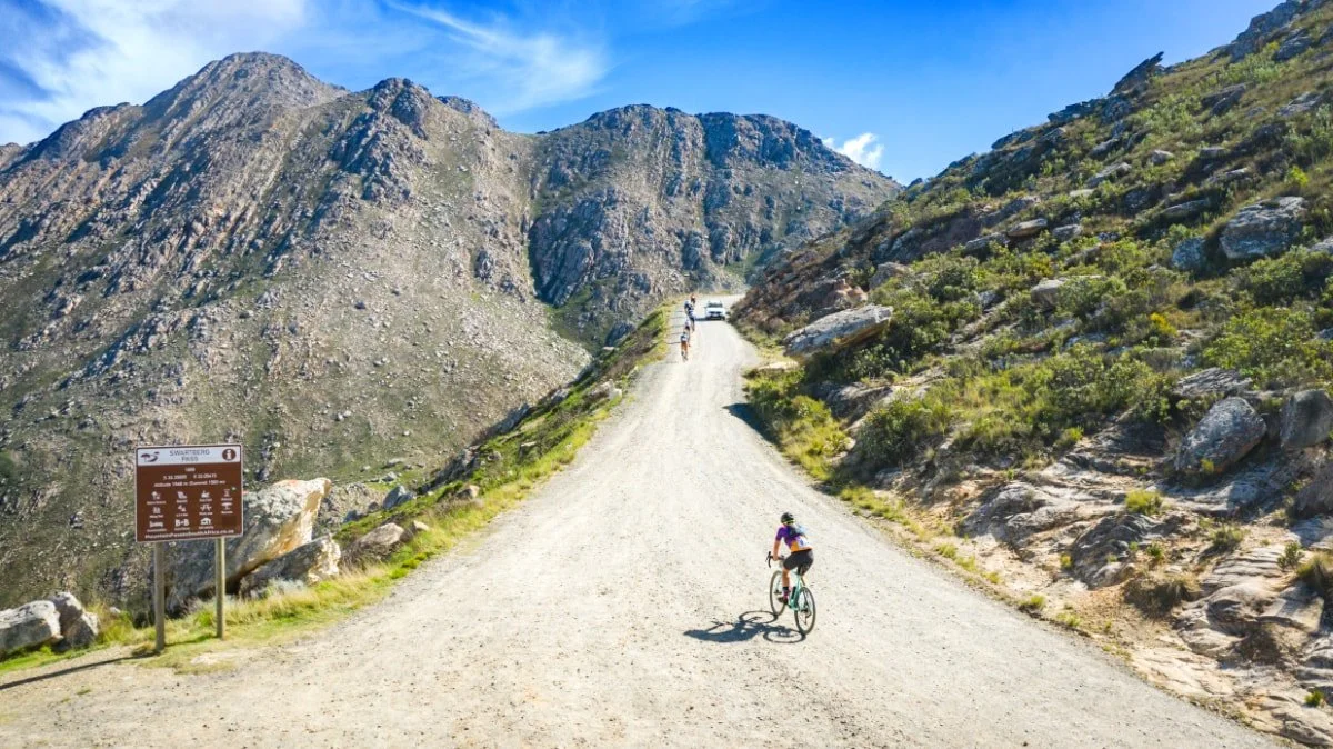 Cyclists riding on a dirt mountain trail with rocky hills and mountains in the background under a blue sky.