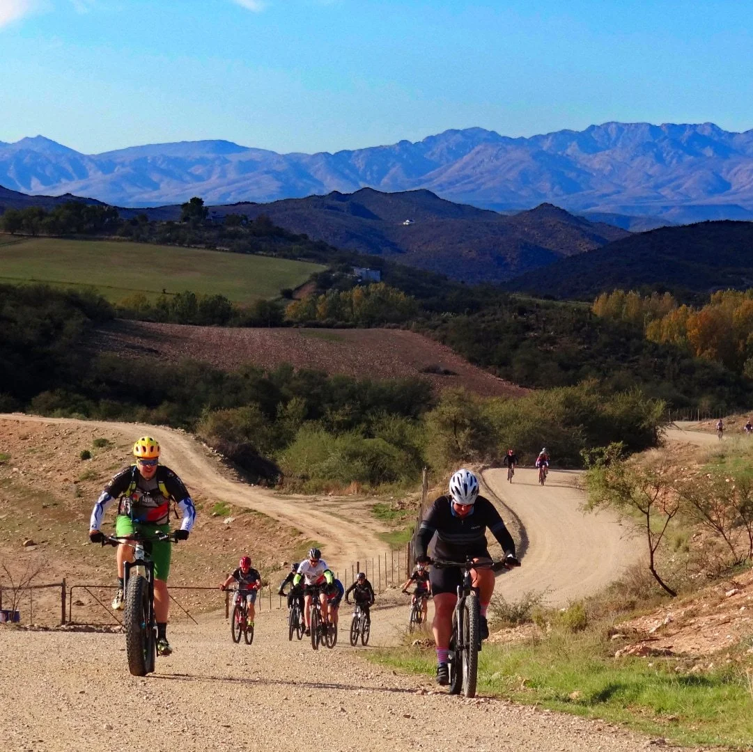 Group of people riding mountain bikes on a dirt trail through hilly landscape with mountains in the background.