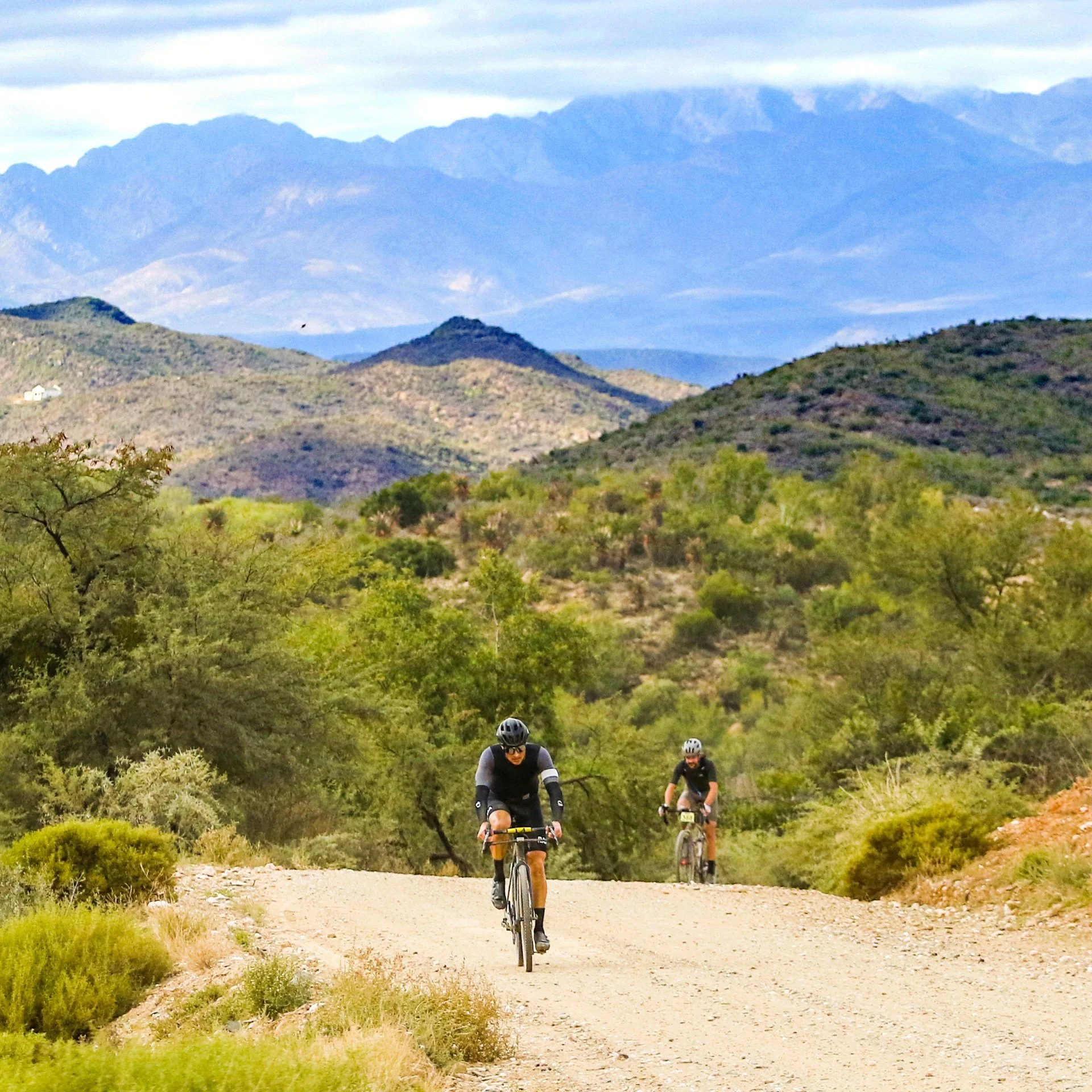 Two cyclists riding on a dirt trail through green shrubbery and trees, with mountains in the background under a partly cloudy sky.