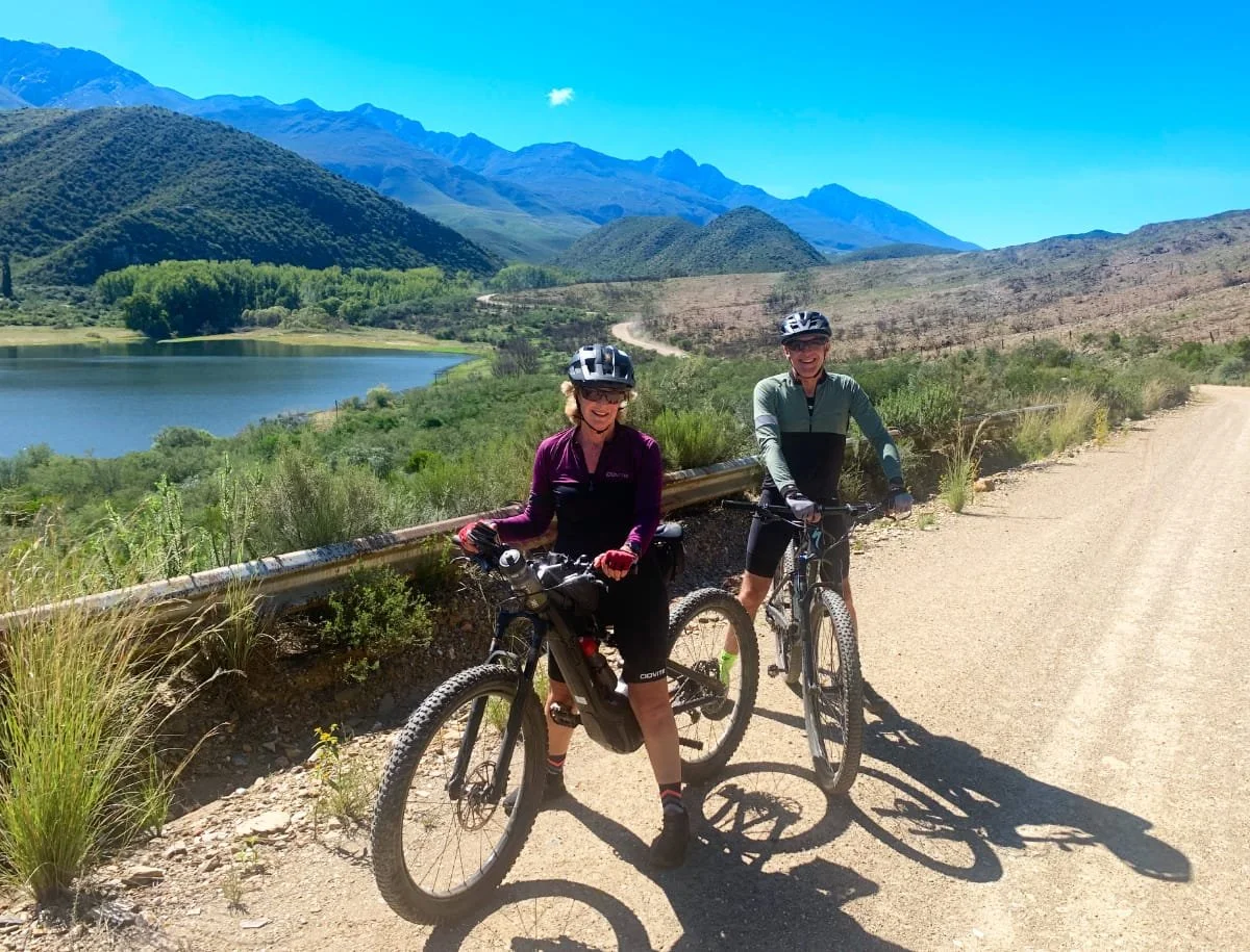 Two women in hiking gear with helmets riding mountain bikes on a dirt trail near a lake, with mountains in the background under a clear blue sky.