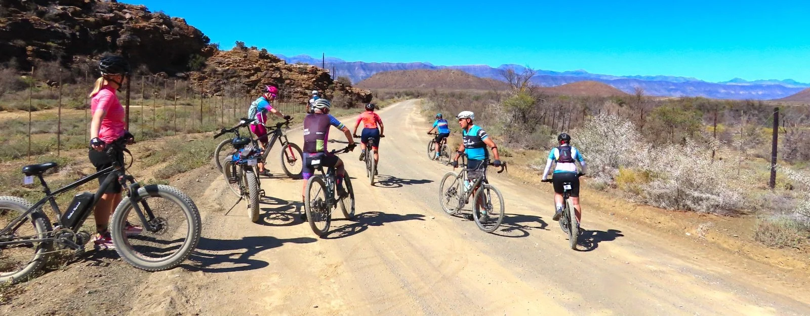 A group of cyclists on a dirt trail in a desert landscape with mountains in the background.