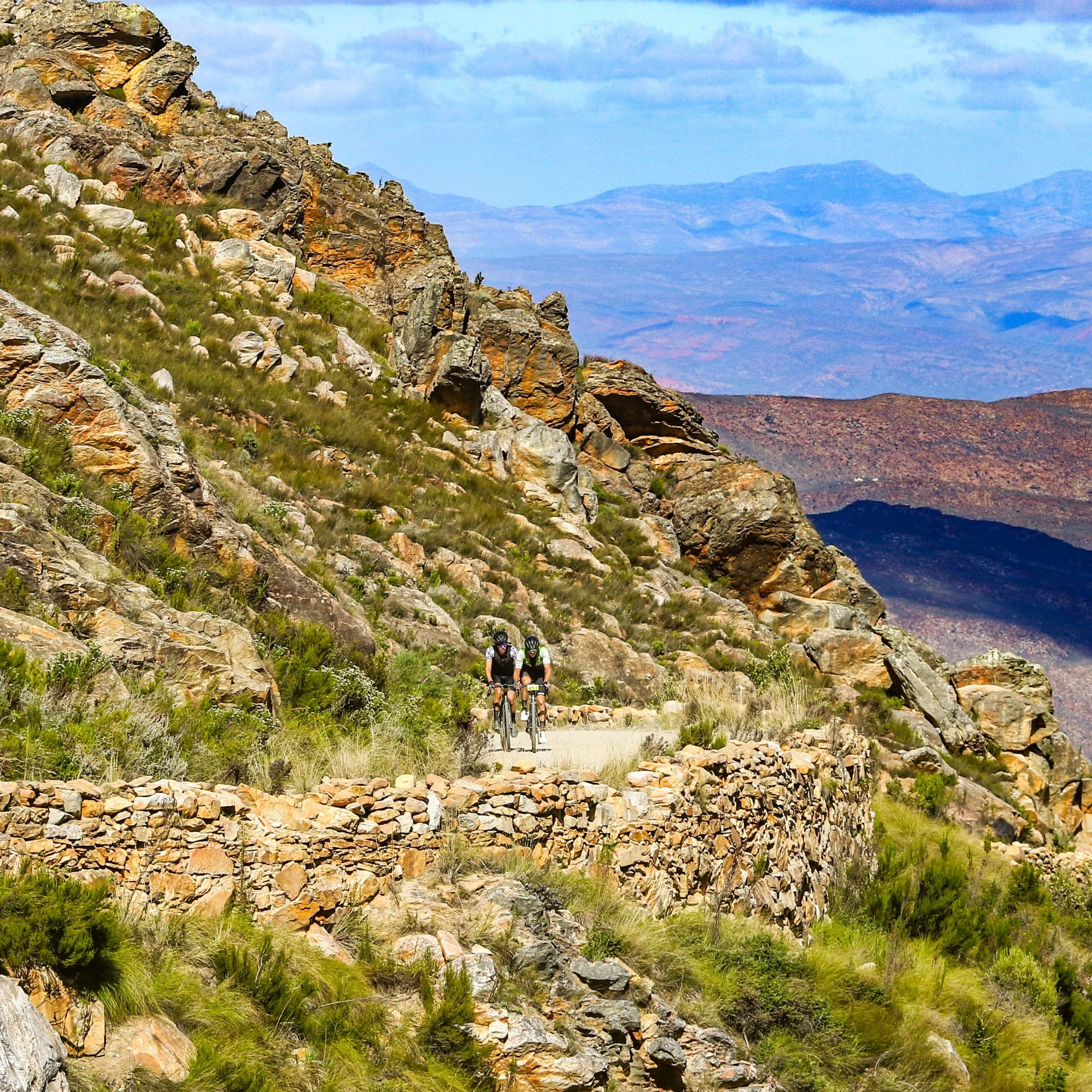Two cyclists riding on a narrow mountain trail with rocky terrain, surrounded by green vegetation and rocky hills under a partly cloudy sky.