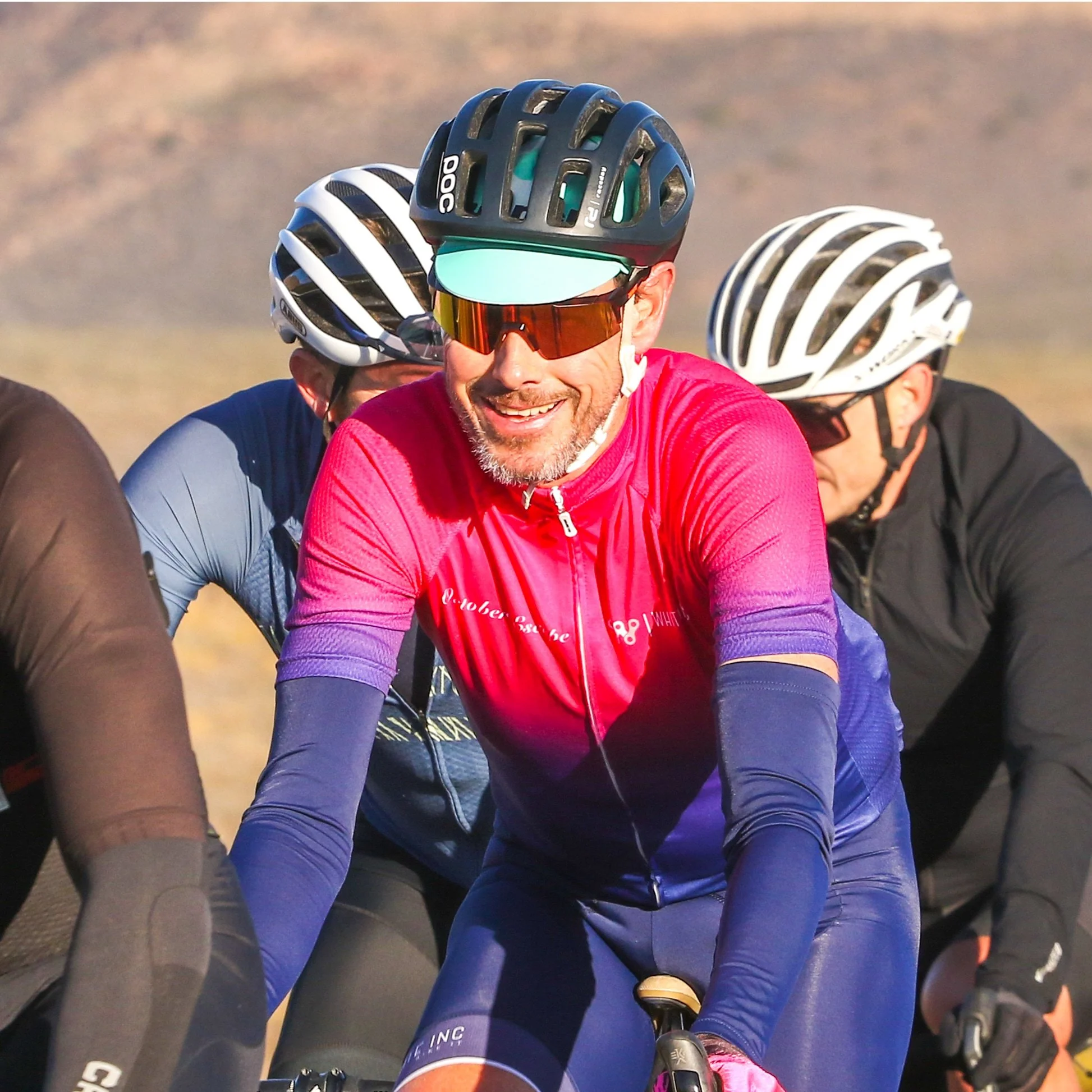 Group of cyclists wearing helmets and sunglasses riding outdoors in sunny weather.