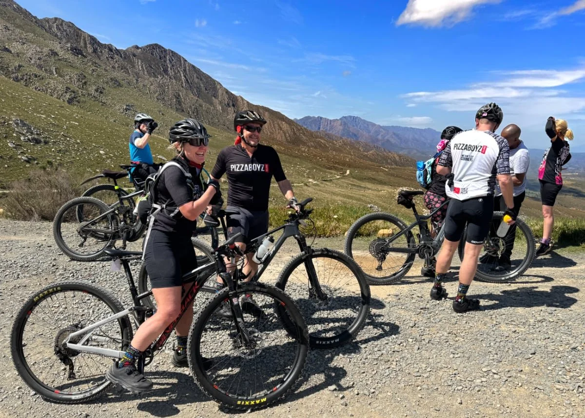 Group of cyclists taking a break on a mountain trail with green rolling hills and mountains in the background on a sunny day.