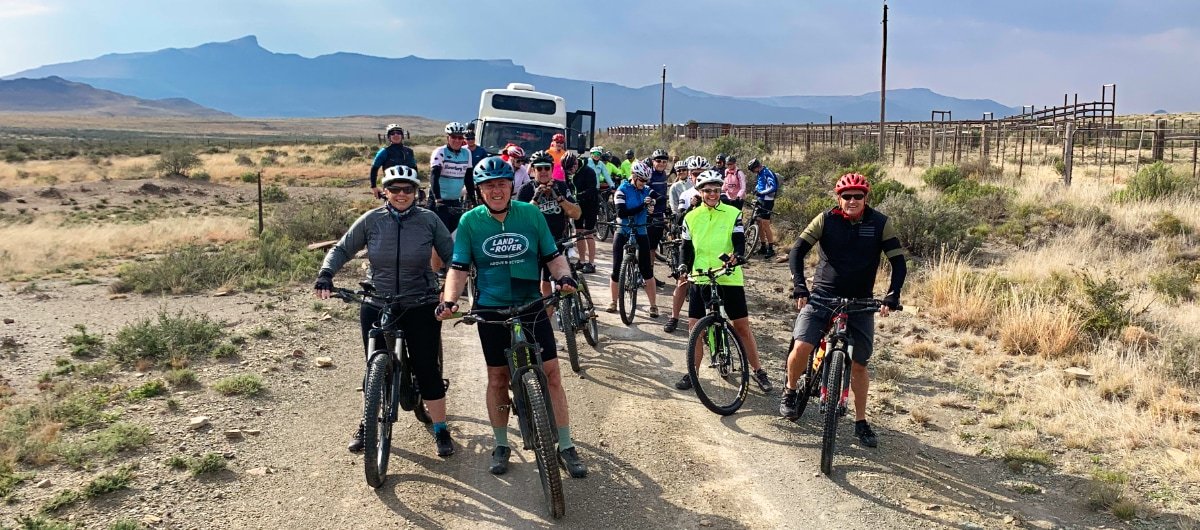 Group of people with bicycles in a desert landscape, mountains in the background, and a bus parked on a dirt road.