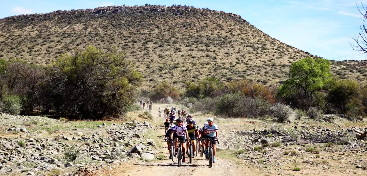 Group of cyclists riding on a dusty trail through a semi-arid landscape with sparse vegetation and a large hill in the background.