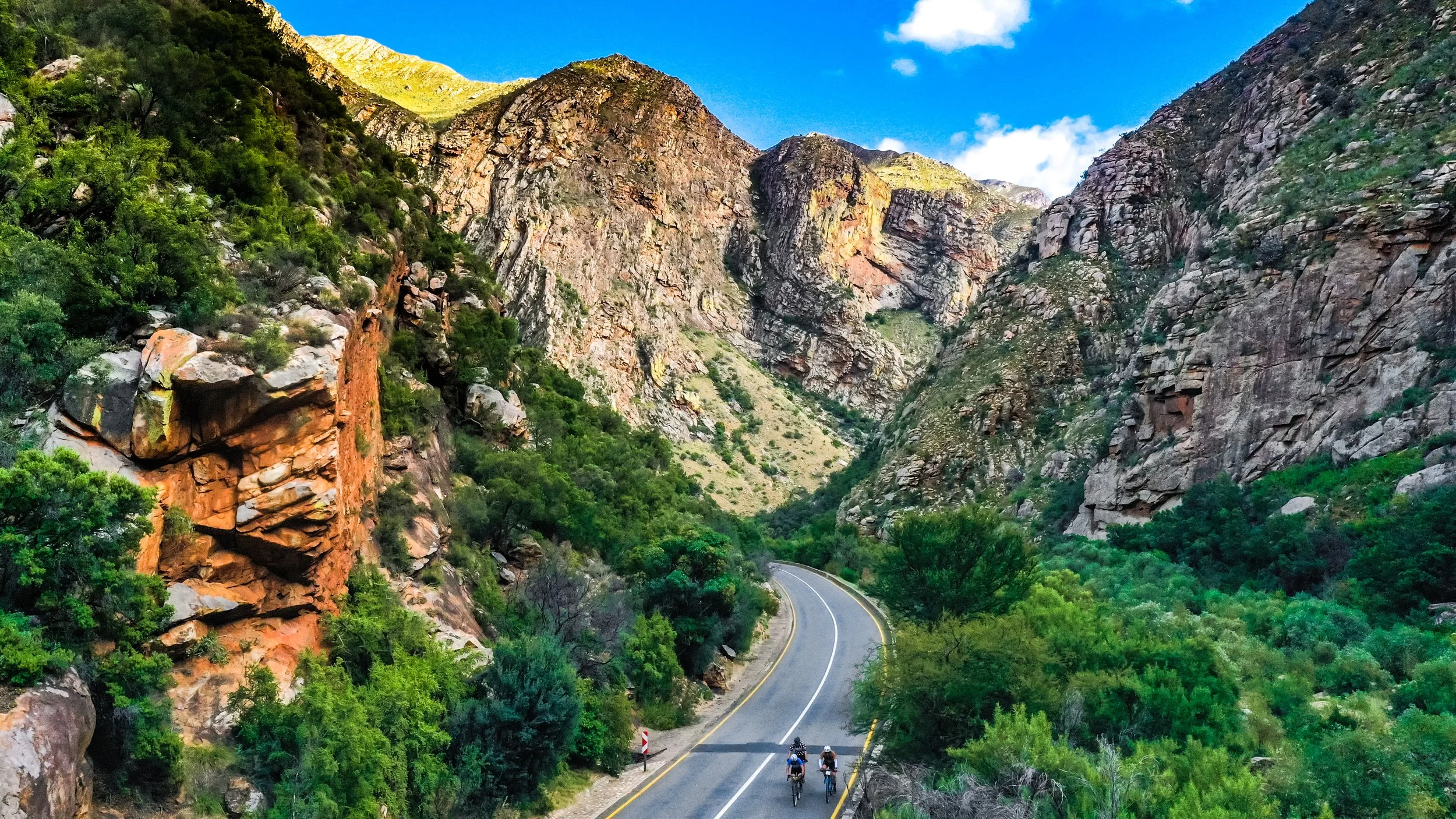 Two cyclists riding on a mountain road surrounded by rocky cliffs and lush green vegetation, with blue sky and white clouds overhead.