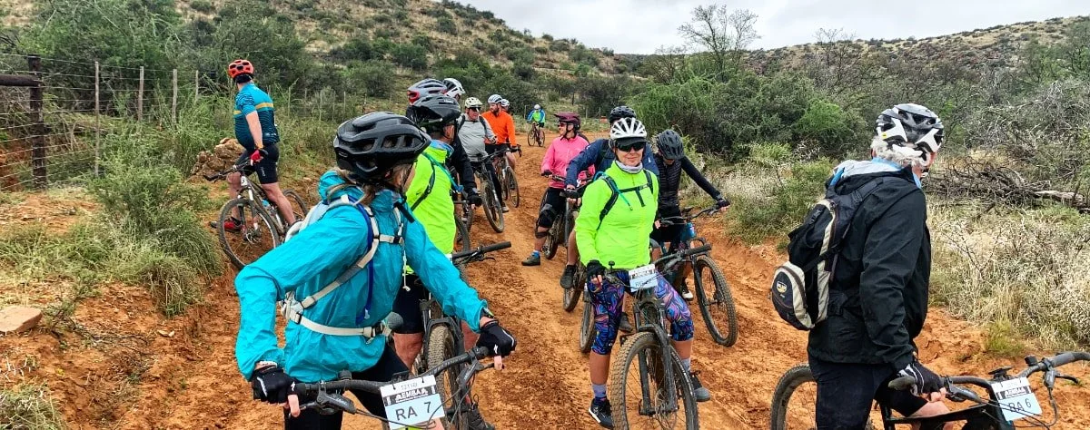 Group of people wearing helmets and colorful outdoor clothing on mountain bikes on a dirt trail with green shrubs and trees in a hilly landscape.
