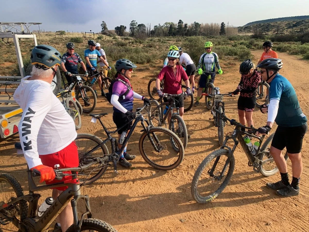 Group of people with mountain bikes gathered outdoors on a dirt trail, some adjusting bikes and others chatting, with a desert landscape and cloudy sky in the background.