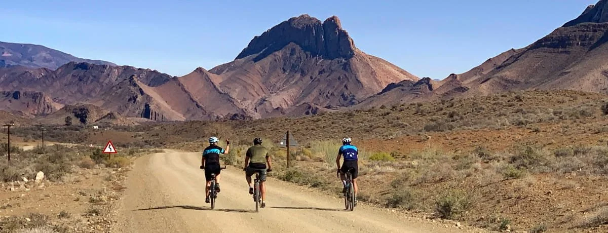Three cyclists riding on a dirt road in a desert landscape with mountains in the background.