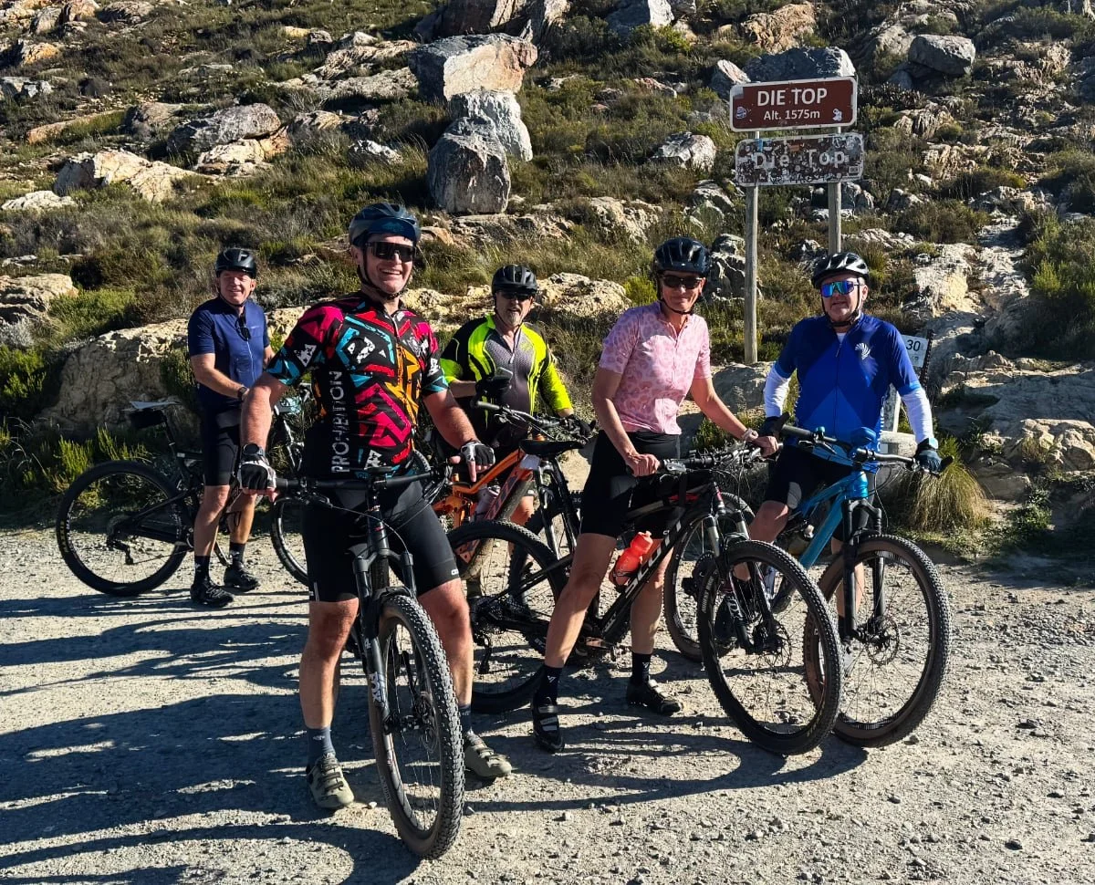 Group of five cyclists with mountain bikes at Die Top, altitude 1575 meters, on a rocky trail with sparse vegetation and a signpost.