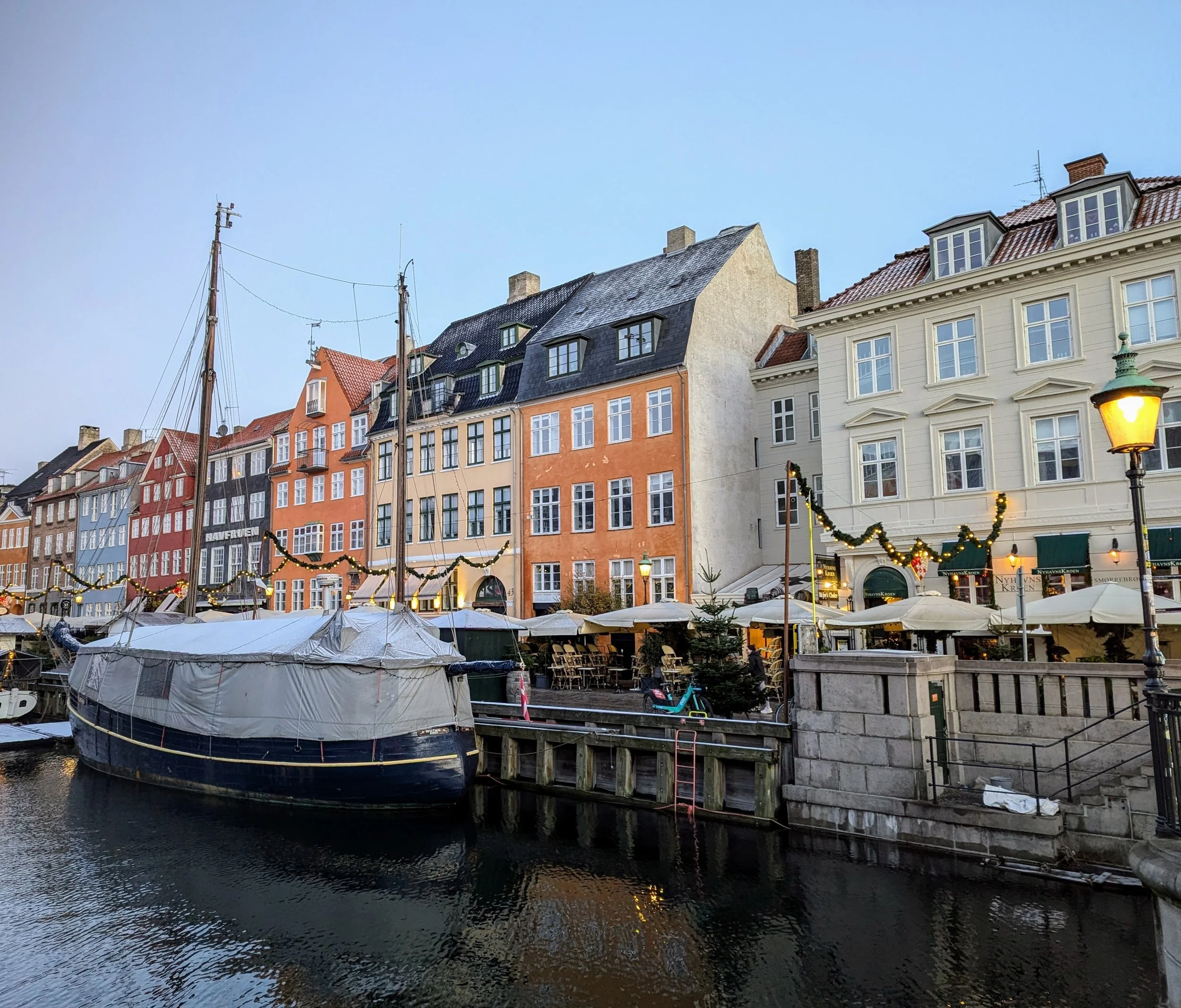Colorful European harbor with a large boat docked near a stone pier, overlooking a row of historic buildings decorated with holiday garlands and lights, under a clear blue sky.