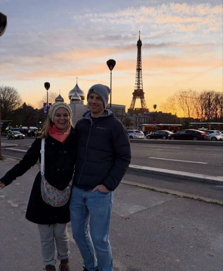 Two people smile and pose for a photo in front of the Eiffel Tower during sunset in Paris.