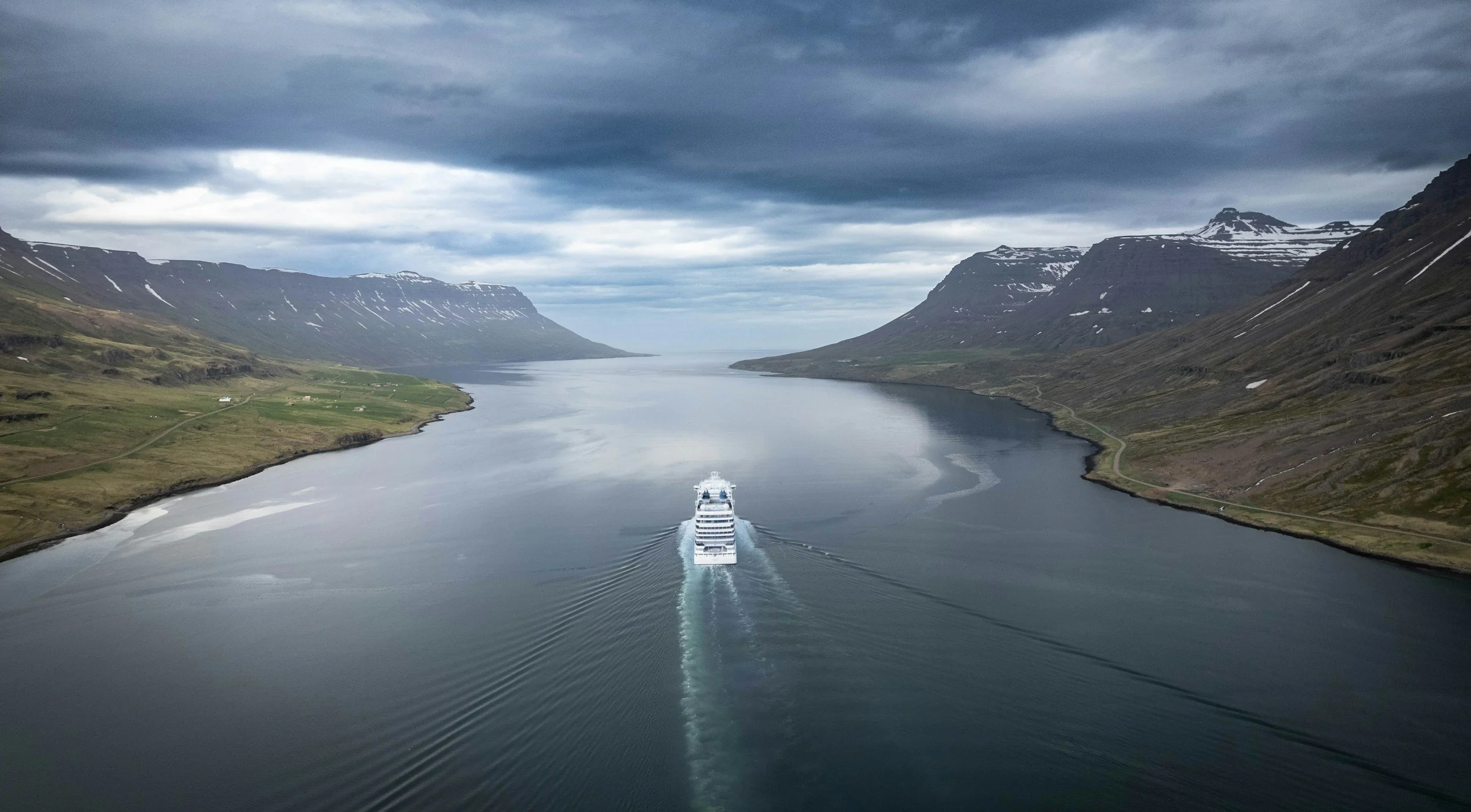 A large white cruise ship sailing through a narrow fjord surrounded by green mountains with patches of snow, under a cloudy sky.