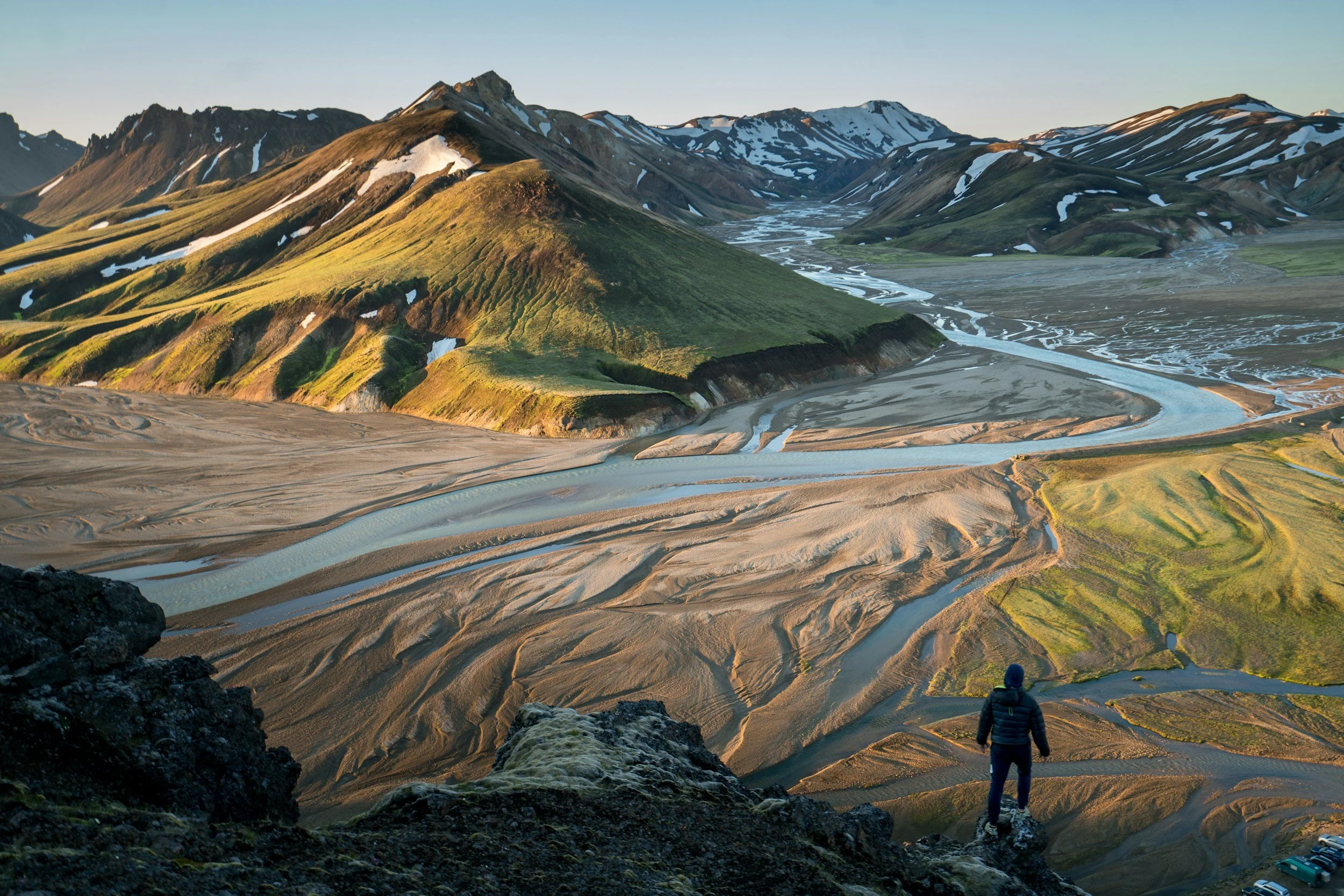 A hiker stands on a rocky ridge overlooking a winding river valley surrounded by mountains with patches of snow and green vegetation.