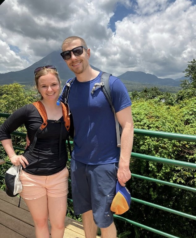 A smiling man and woman standing together outdoors with lush green trees and mountains in the background. They are dressed for hiking, carrying backpacks, and wearing sunglasses.
