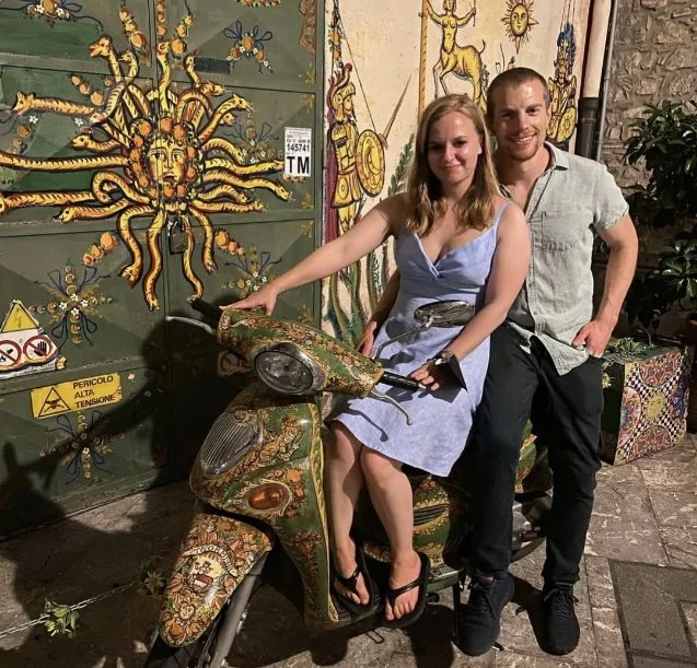 A woman sitting on a decorated scooter with a man standing behind her. They are in front of a colorful wall with artwork, including a large sun face and other decorative elements. The woman is pointing at the scooter, and both are smiling.