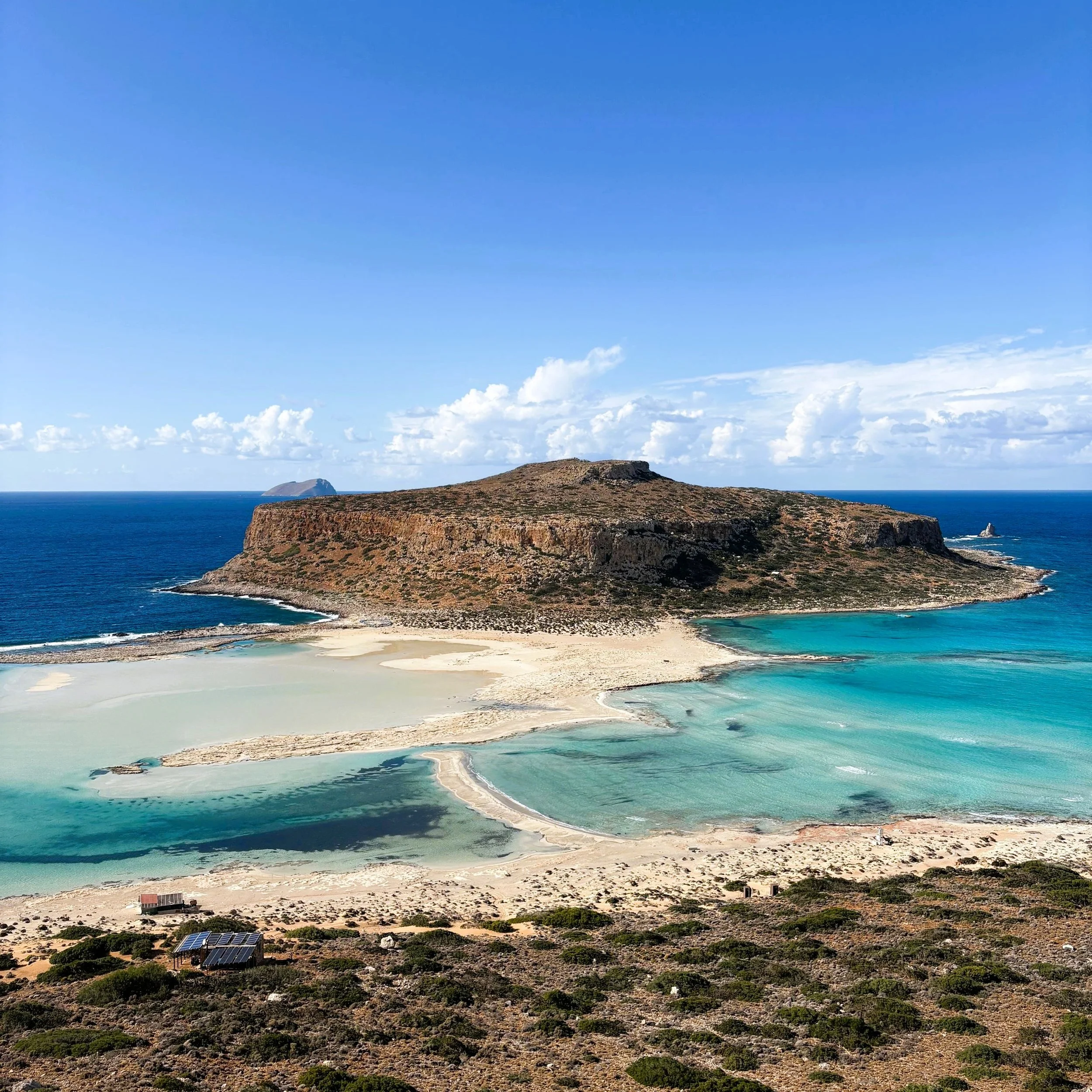 A scenic view of a coastal landscape featuring a large rocky hill surrounded by a lagoon with turquoise waters and sandy shores, under a blue sky with scattered clouds.