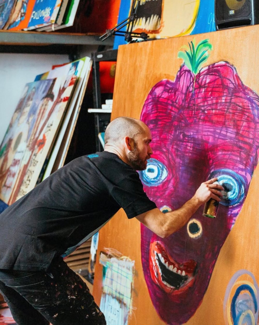 An artist with a shaved head and beard creating a colorful painting of a face with exaggerated features on a large canvas in an art studio.