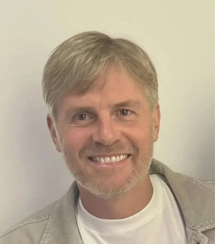 A smiling man with blonde hair and a beard, wearing a white t-shirt and a light-colored jacket, standing against a plain light-colored wall.