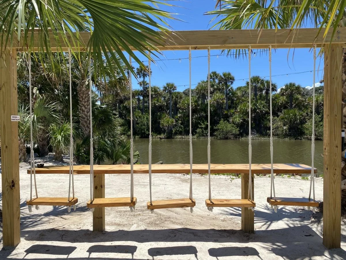 Four wooden swings hanging from a sturdy wooden frame with ropes, set on a sandy area near a river with lush green trees and palm trees in the background under a clear blue sky.