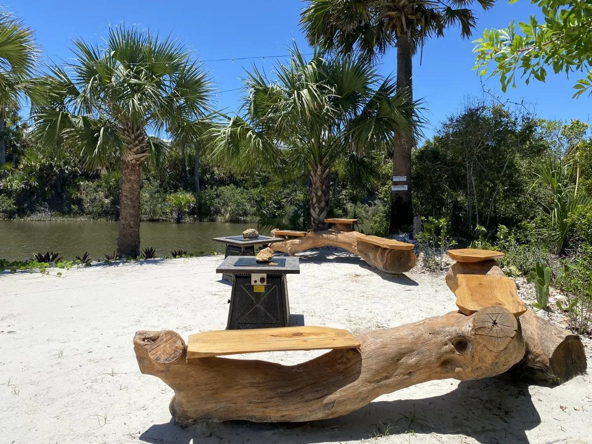 Wooden benches and tables on white sand near palm trees by a body of water under a clear blue sky.