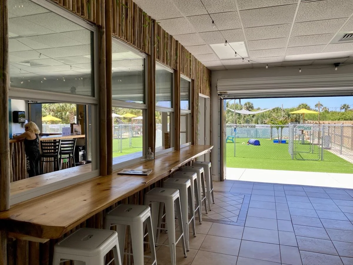 Indoor area with a wooden bar-style counter, several white stools, large windows showing an outdoor playground area with green grass, yellow umbrellas, and play equipment.