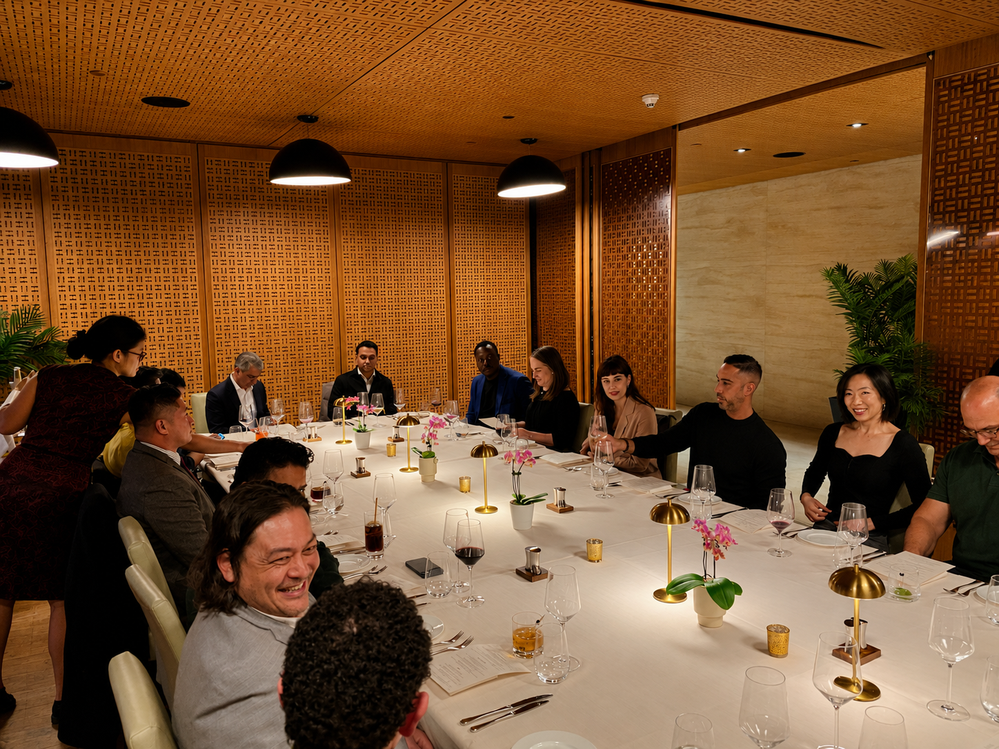A diverse group of seven people in formal attire enjoying a dinner conversation at a round table with candles, wine glasses, and a floral centerpiece in an elegant dining room.