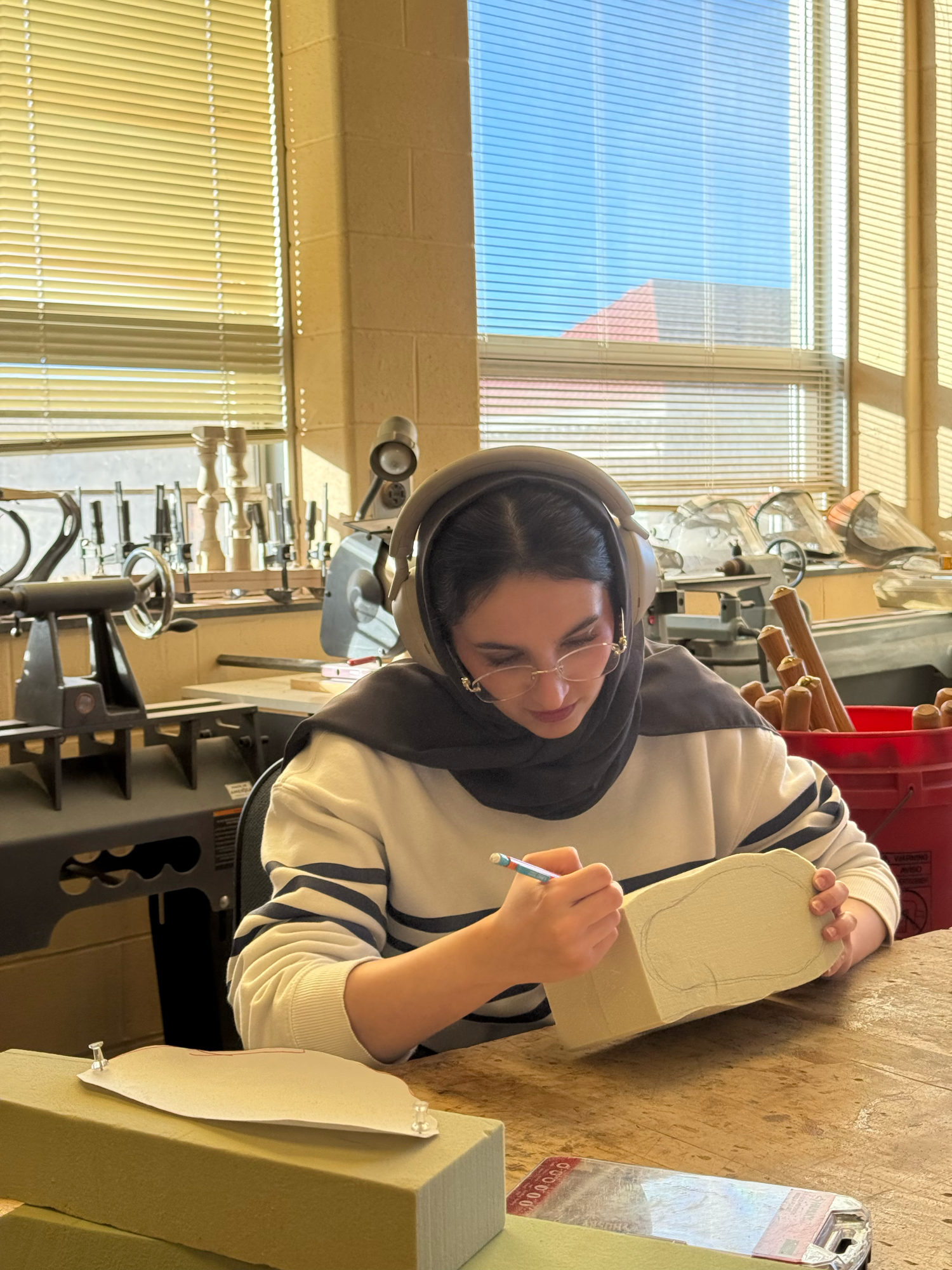 A young woman wearing glasses, a black hijab, and headphones is working on a sculpture of a face in a workshop with large windows and tools.