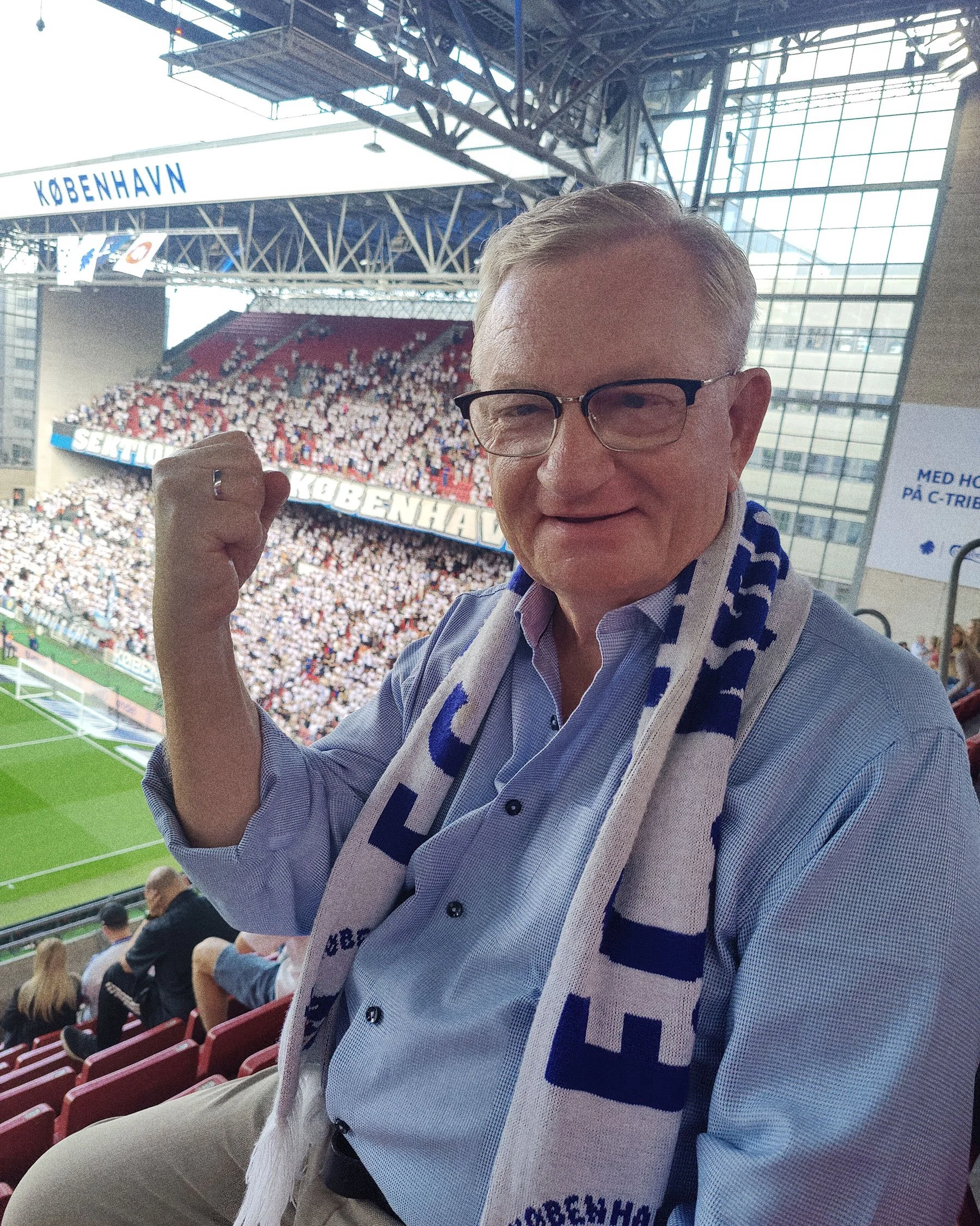 Smiling man wearing glasses and a blue shirt with a blue and white scarf, sitting in a stadium filled with fans.