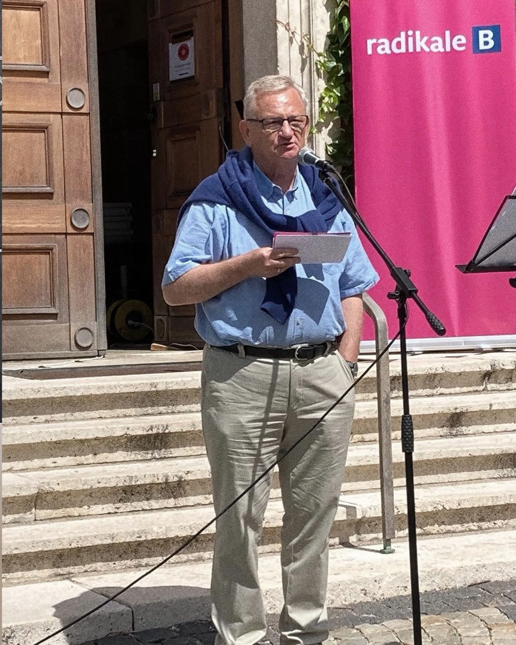 A man in a blue shirt and beige pants speaking into a microphone at an outdoor event, standing on steps in front of a building with a pink banner displaying the word 'radikale' and a blue 'B' logo.