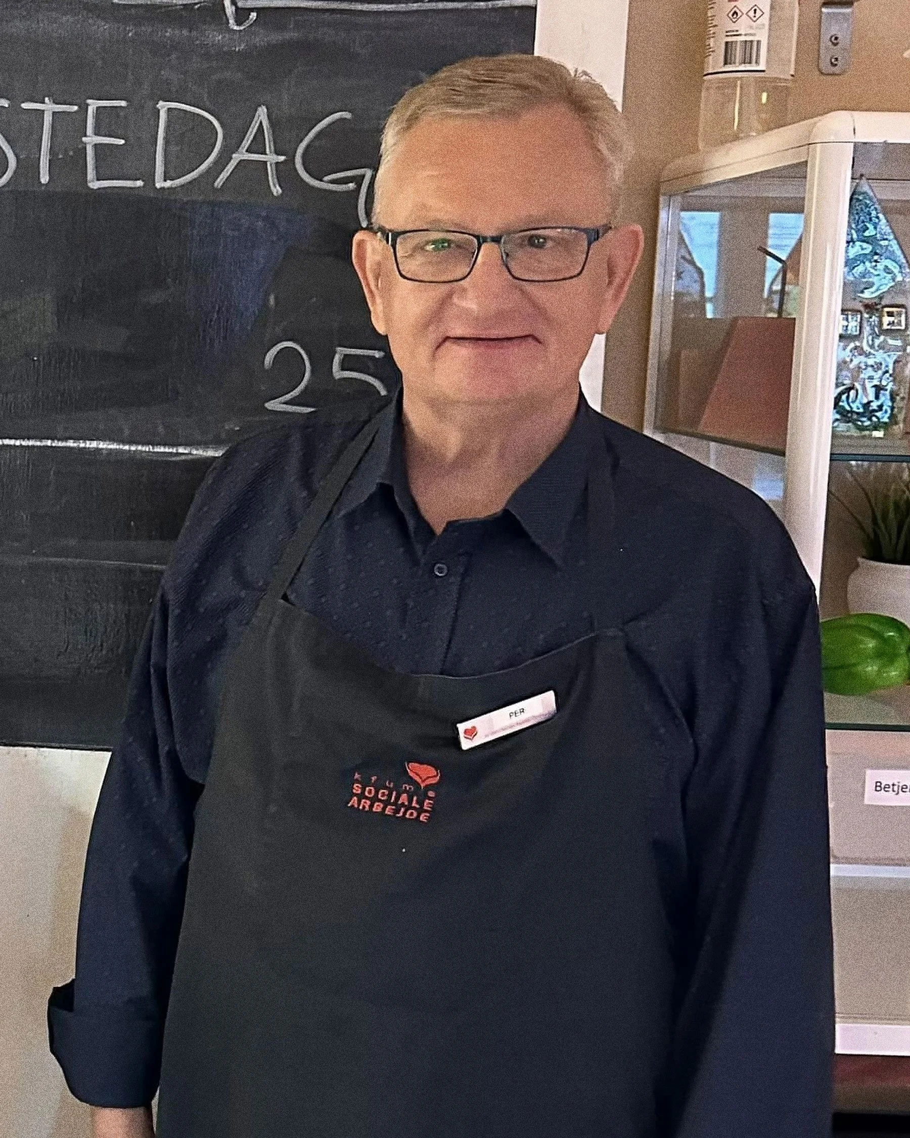 A man wearing glasses and a black apron with red text, standing inside a shop or café, with a blackboard behind him and a shelf with green peppers and a potted plant to his right.