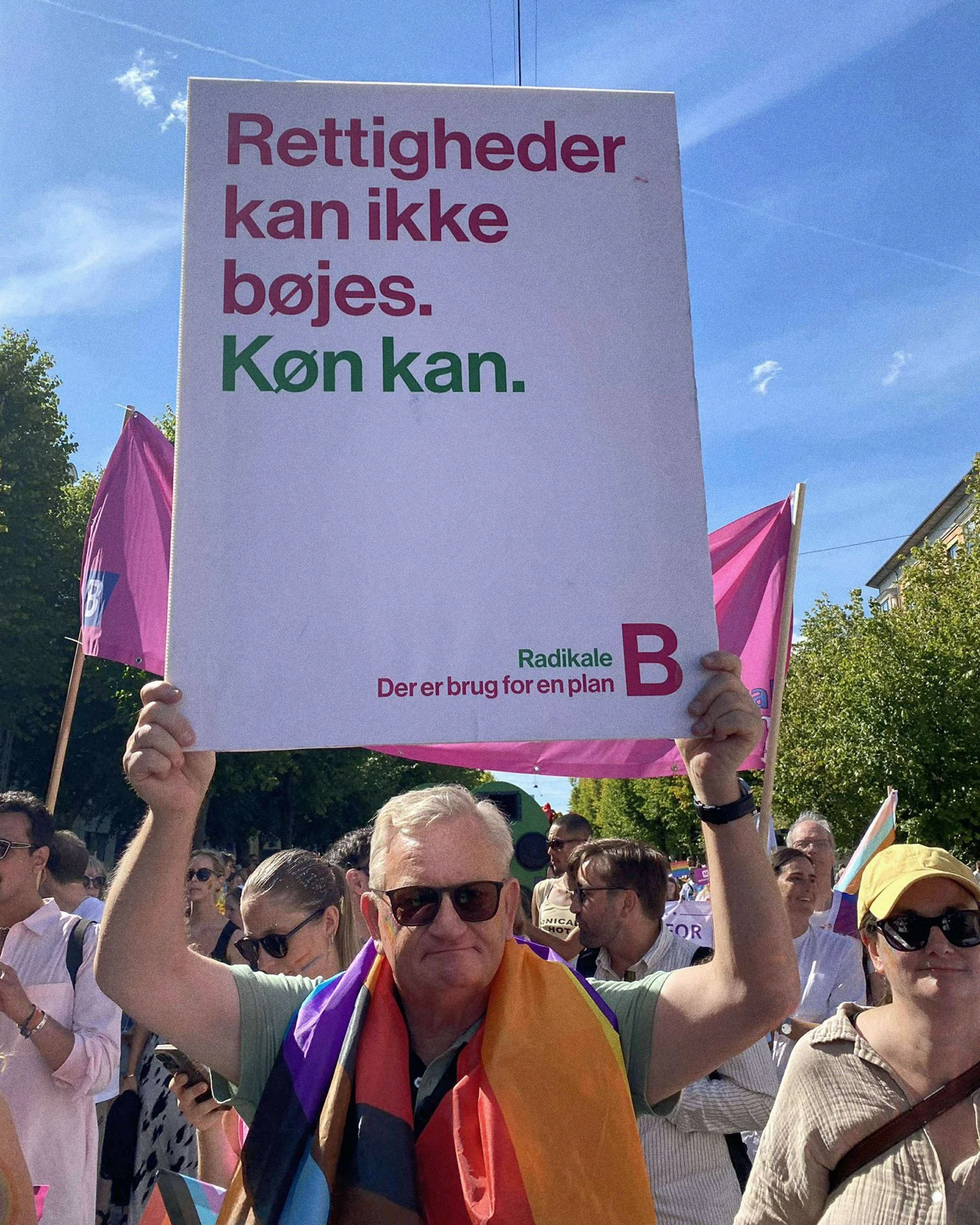 Person holding a protest sign at a rally, with other people and rainbow flags in the background on a sunny day.