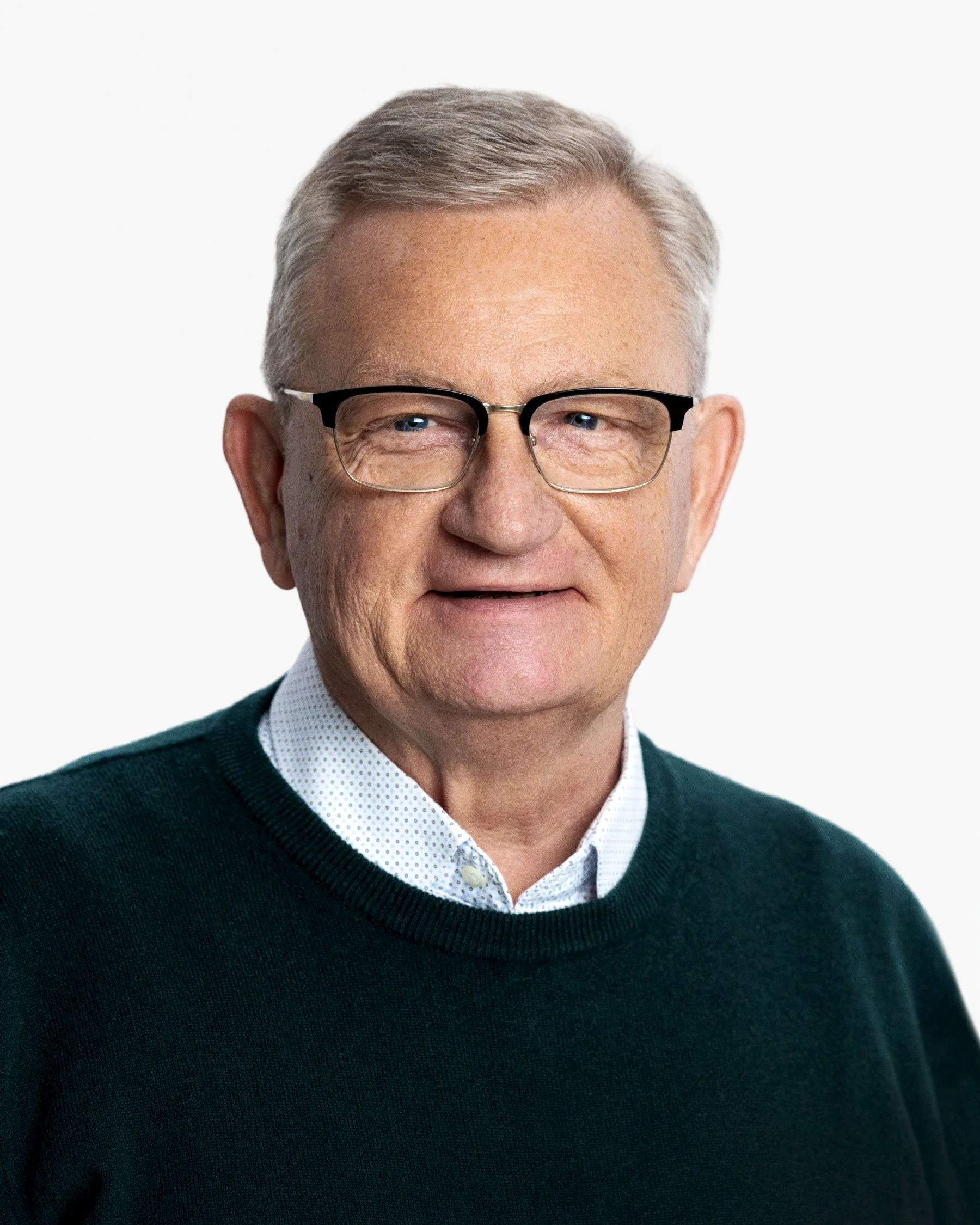 Close-up of an older man wearing glasses, a white collared shirt, and a dark green sweater, smiling against a plain white background.