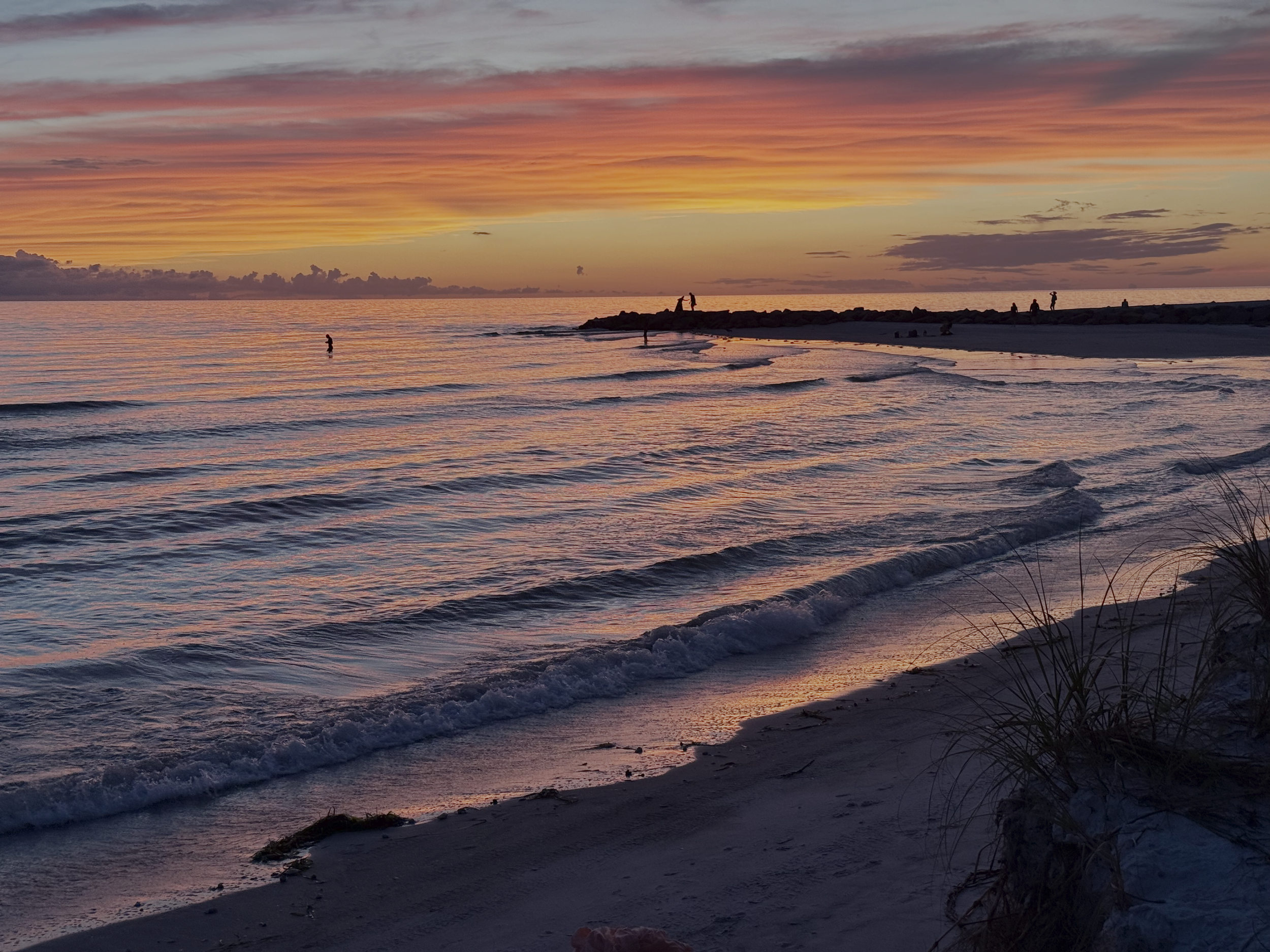 Sunset over the ocean with pink, orange, and purple clouds, gentle waves washing onto the sandy beach, and silhouettes of people on a jetty and shoreline.