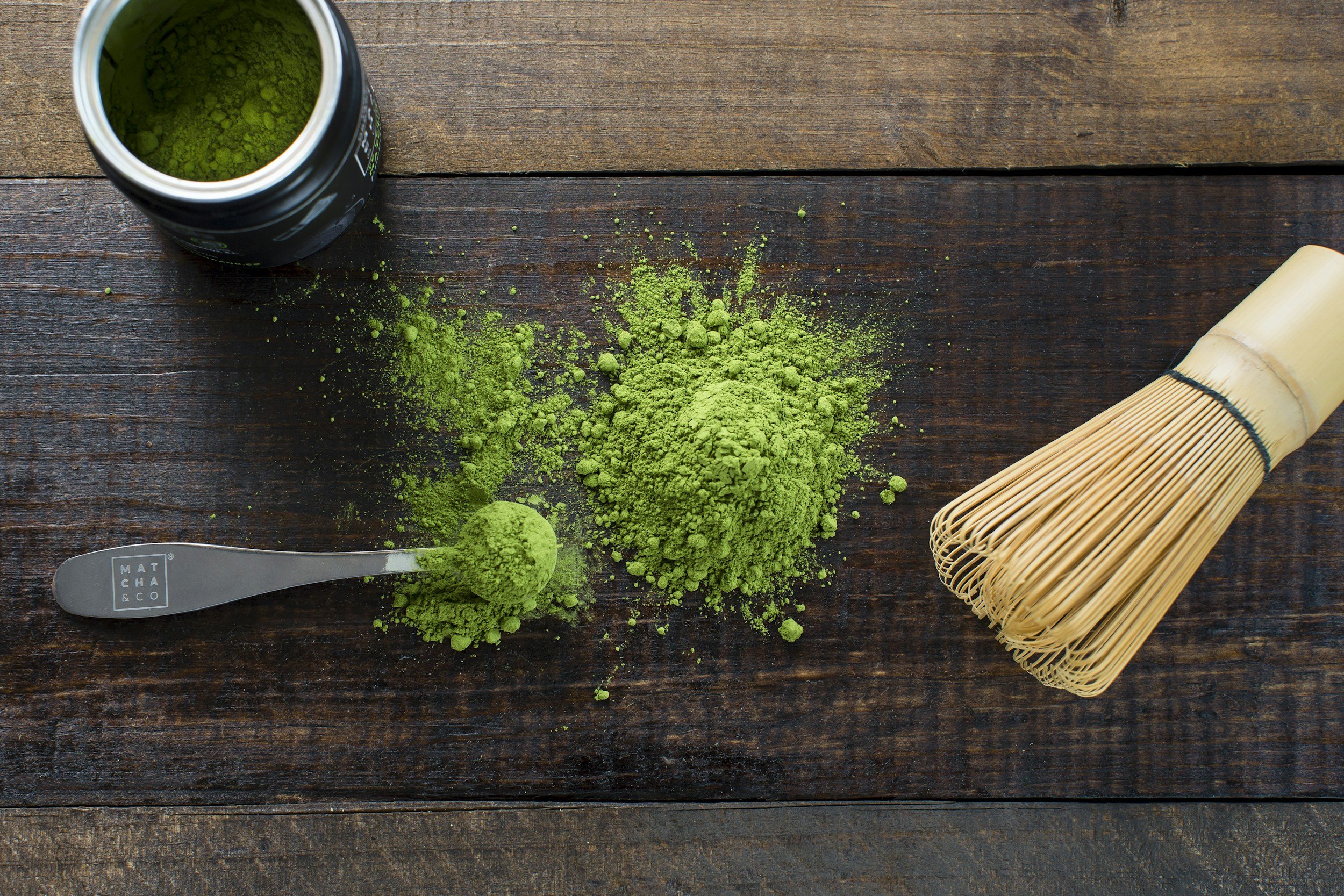 Green matcha powder on a spoon, spilled on a dark wooden surface, with a bamboo whisk and a container of matcha powder.