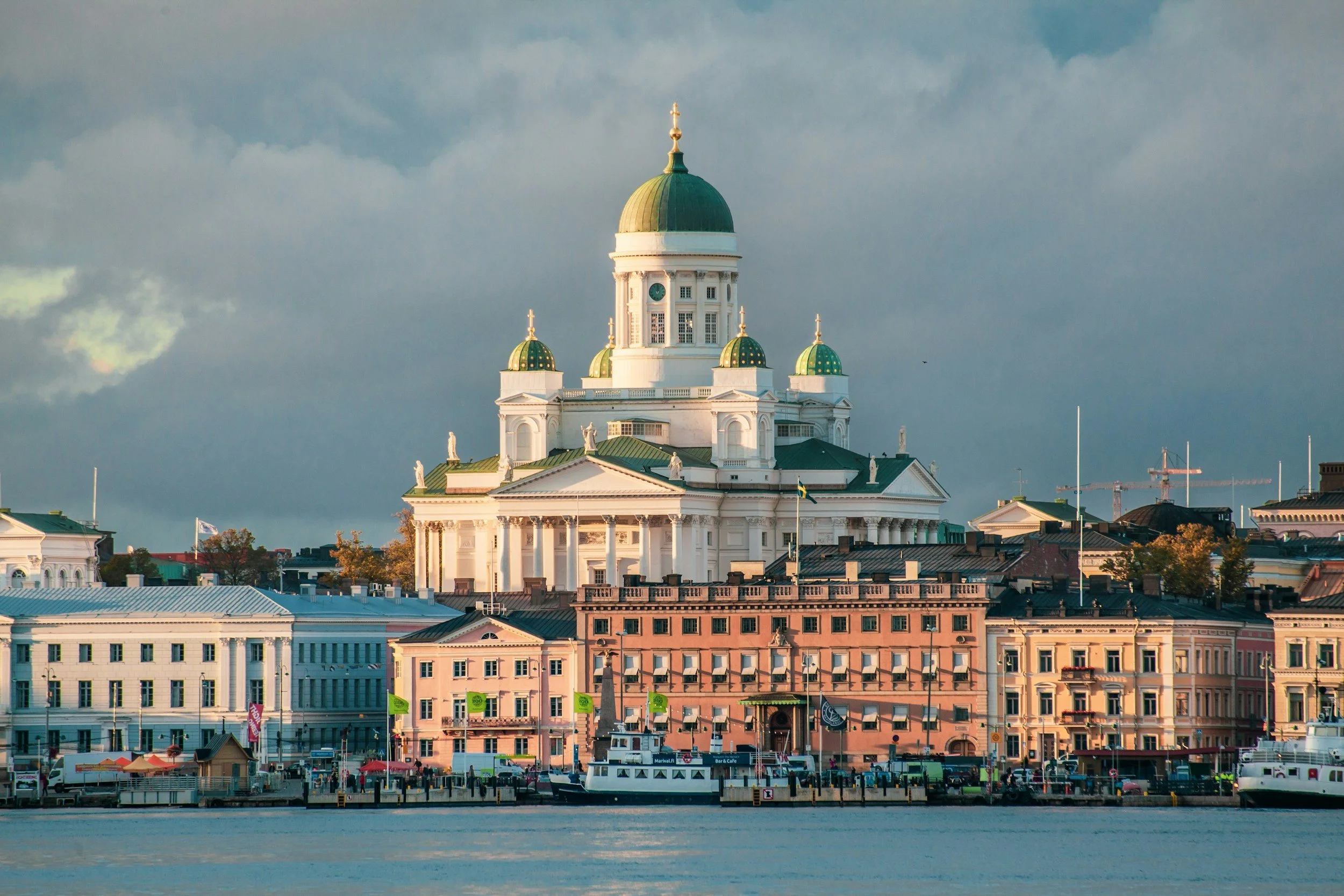 A scenic view of a city waterfront with a large white building featuring a green dome, surrounded by smaller buildings and a docked boat in the foreground.