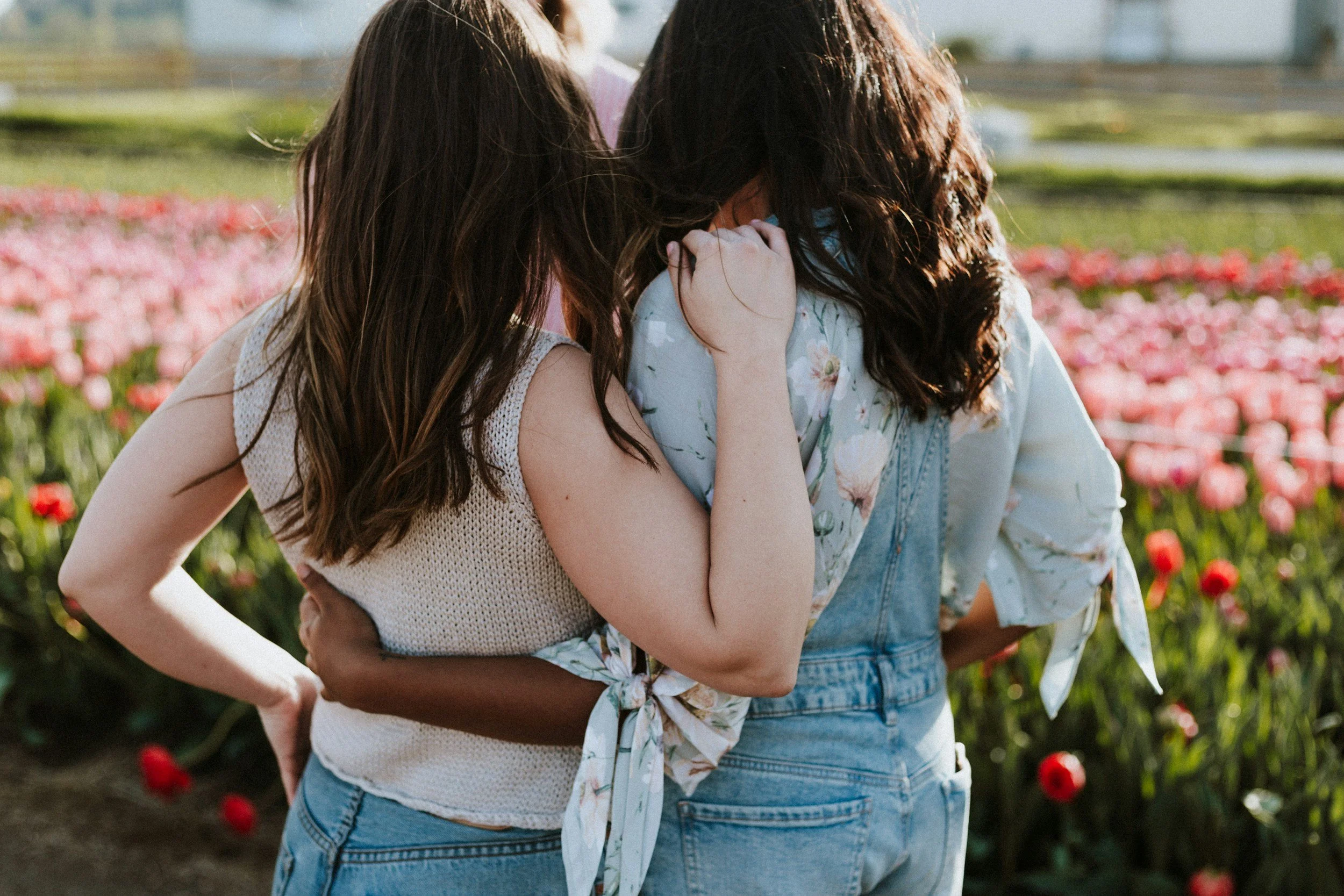 Two women embrace each other in a garden of pink and red flowers, holding each other close.