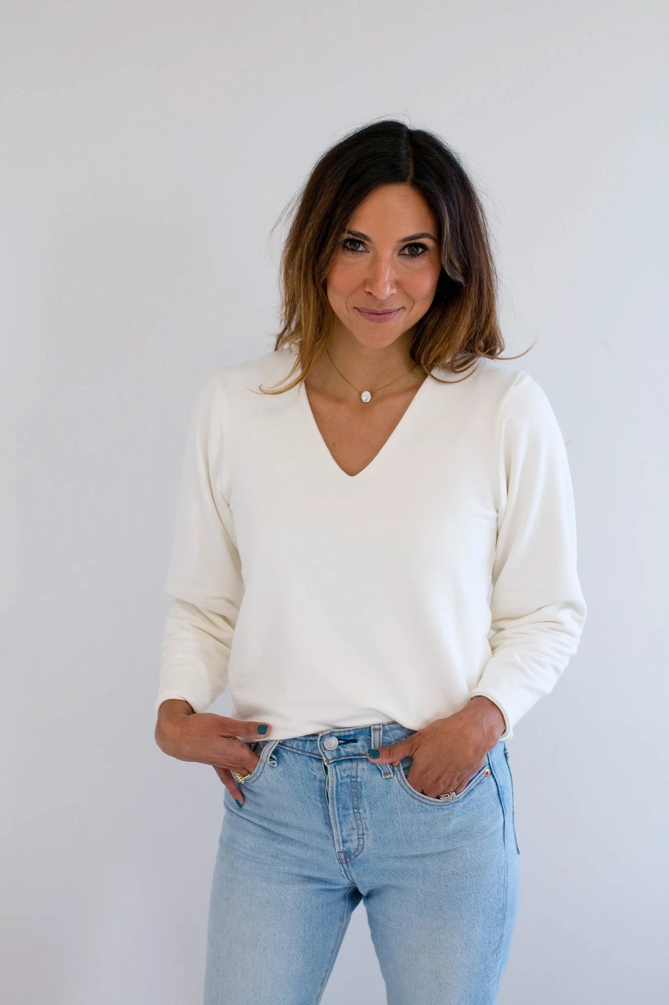 Fertility coach in Edmonton Justine Lapointe with shoulder-length brown hair wearing a white long-sleeve V-neck shirt and light blue jeans, standing against a plain white background.