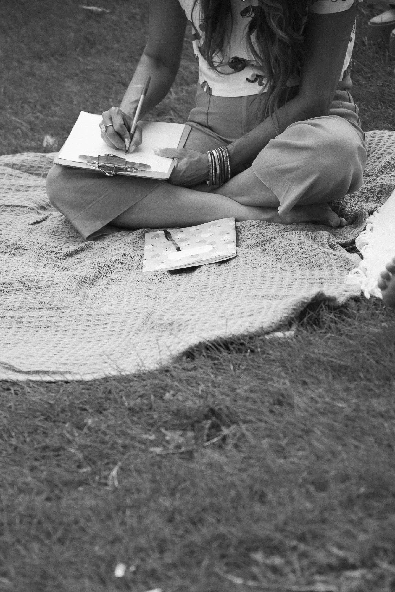 A woman sitting cross-legged on a blanket on the grass, writing in a notebook with a pen. She has long, wavy hair and is wearing bracelets, a short-sleeved top, and loose pants. There is a small notebook and a pen on the blanket.