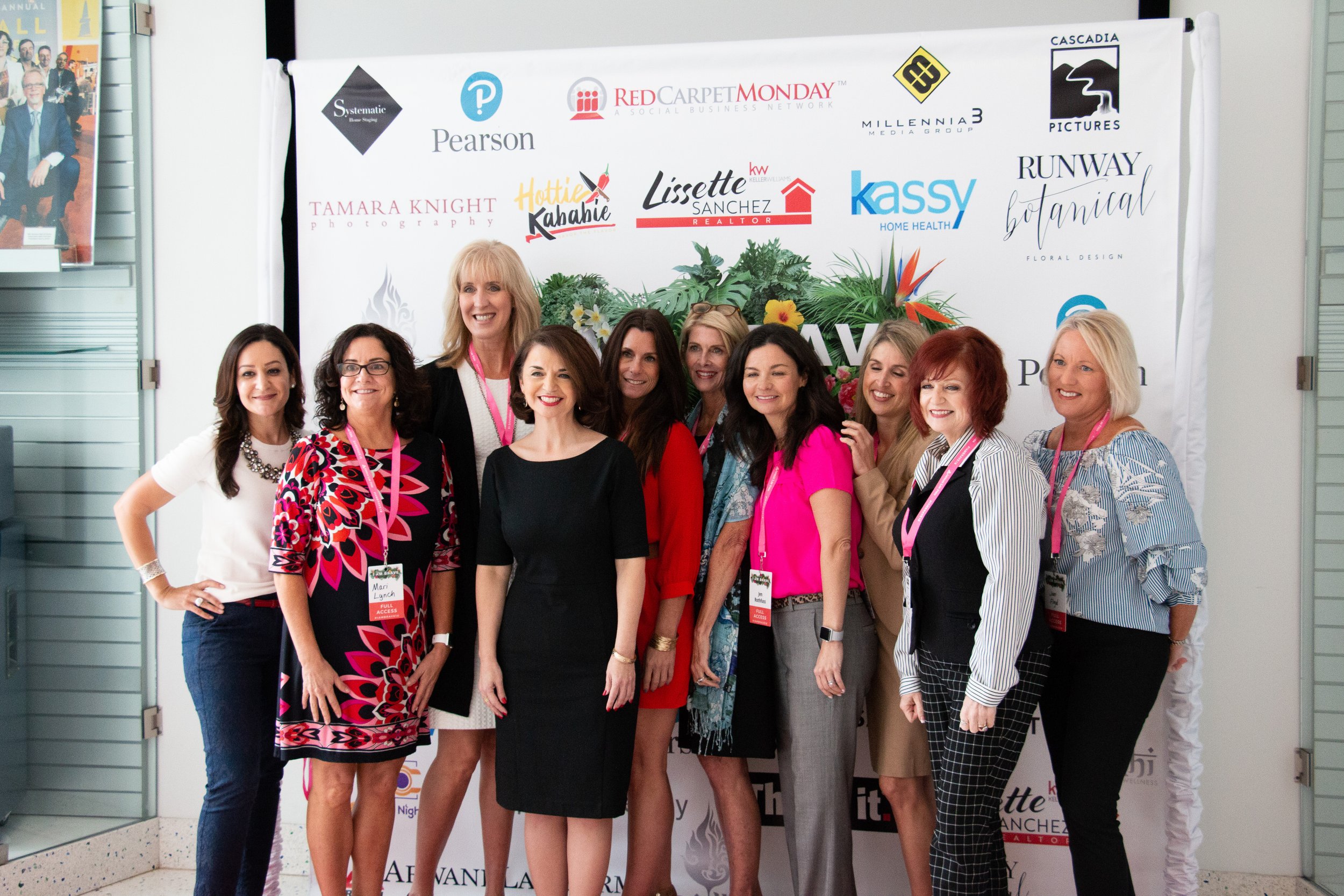 Group of women posing together at a conference or event in front of a backdrop with various brand logos and floral decorations.