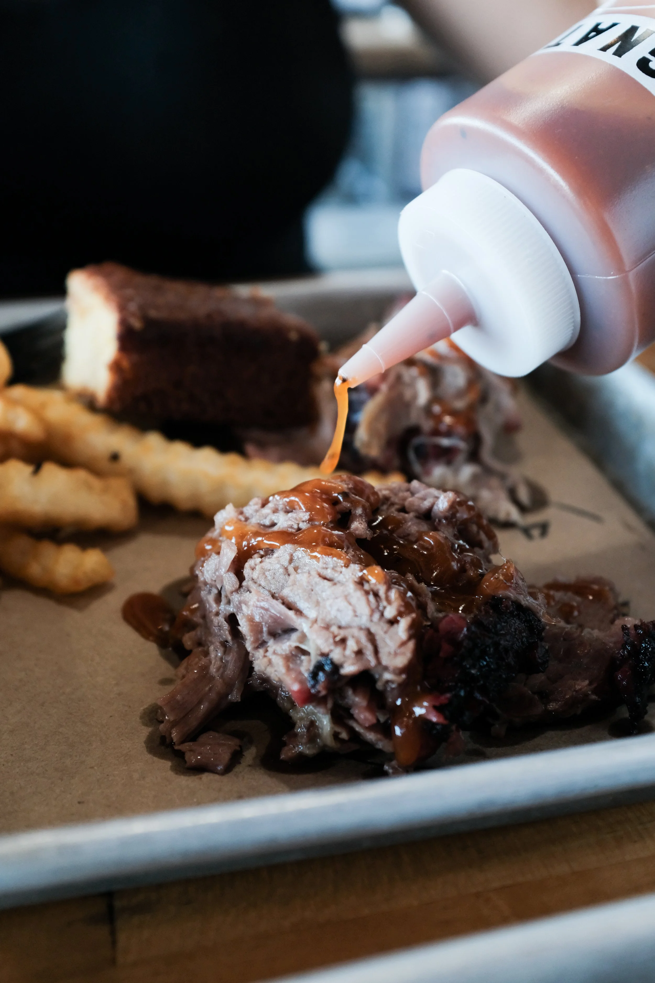 Close-up of barbecue meat with barbecue sauce being squeezed from a bottle, side of French fries, and a slice of chocolate cake on a tray.