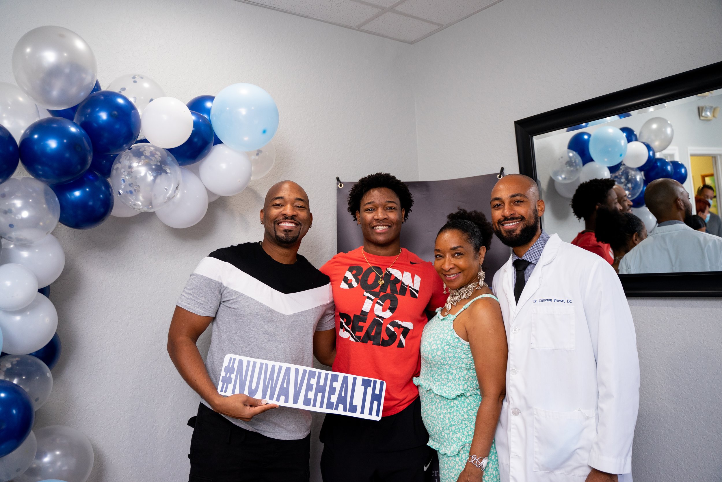 Four people standing indoors at a health awareness event, with a balloon arch in the background and a mirror reflecting the balloons and other attendees. The person on the far left is holding a sign that reads '#NUWAVEHEALTH'. The group includes a ma