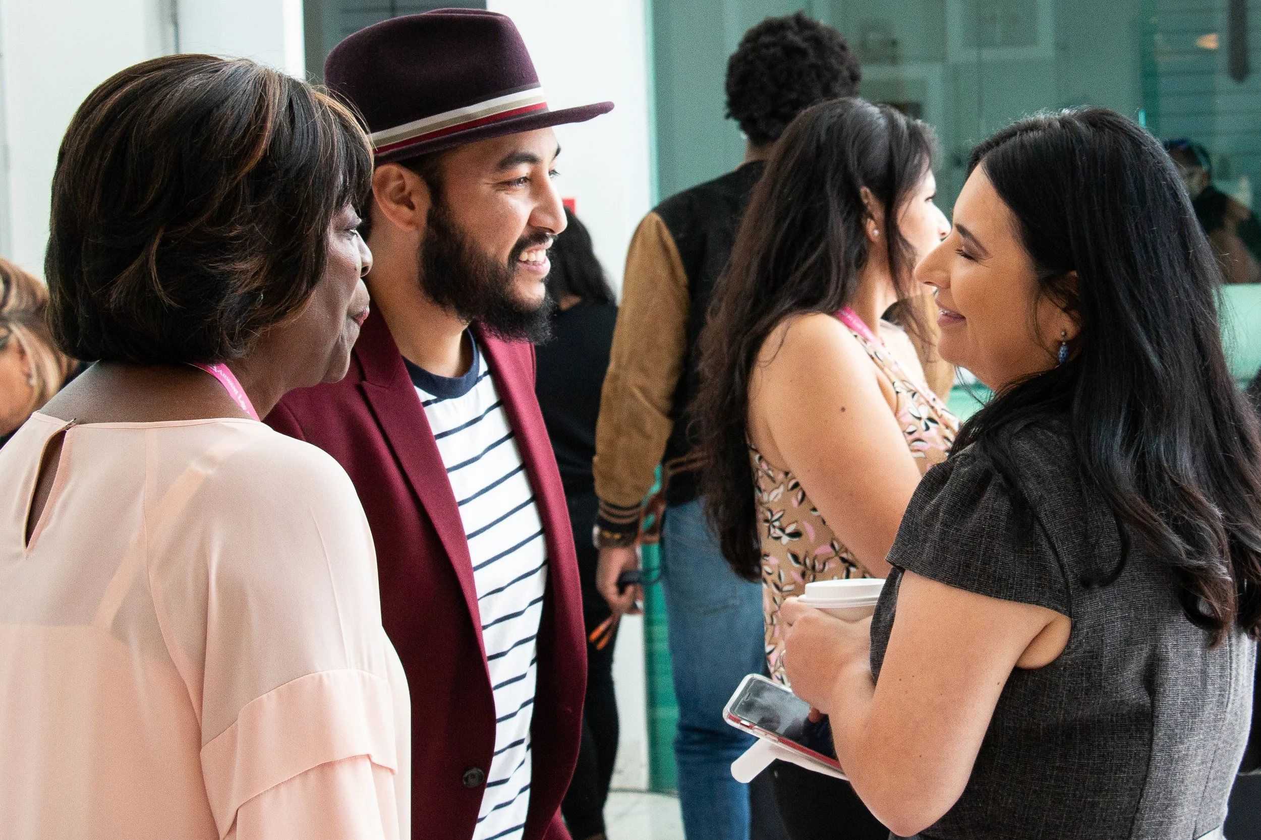 Group of people talking and smiling at an indoor event, diverse in age and ethnicity, with some holding coffee cups and smartphones.