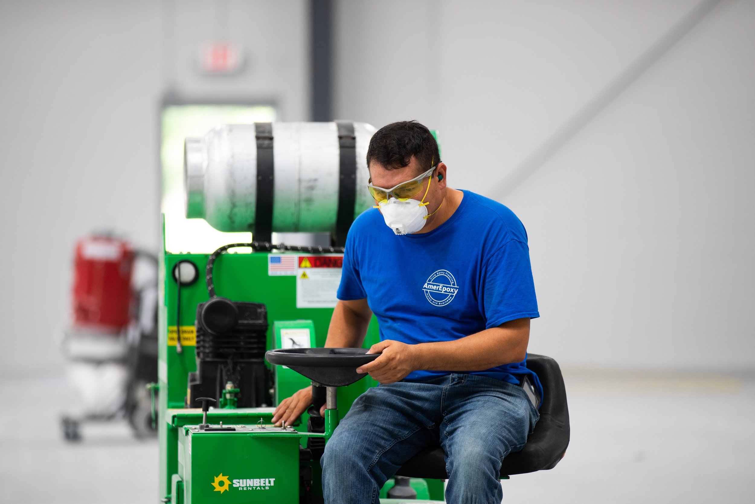 A man wearing safety glasses and a face mask working with a green concrete cutting machine in an industrial setting.