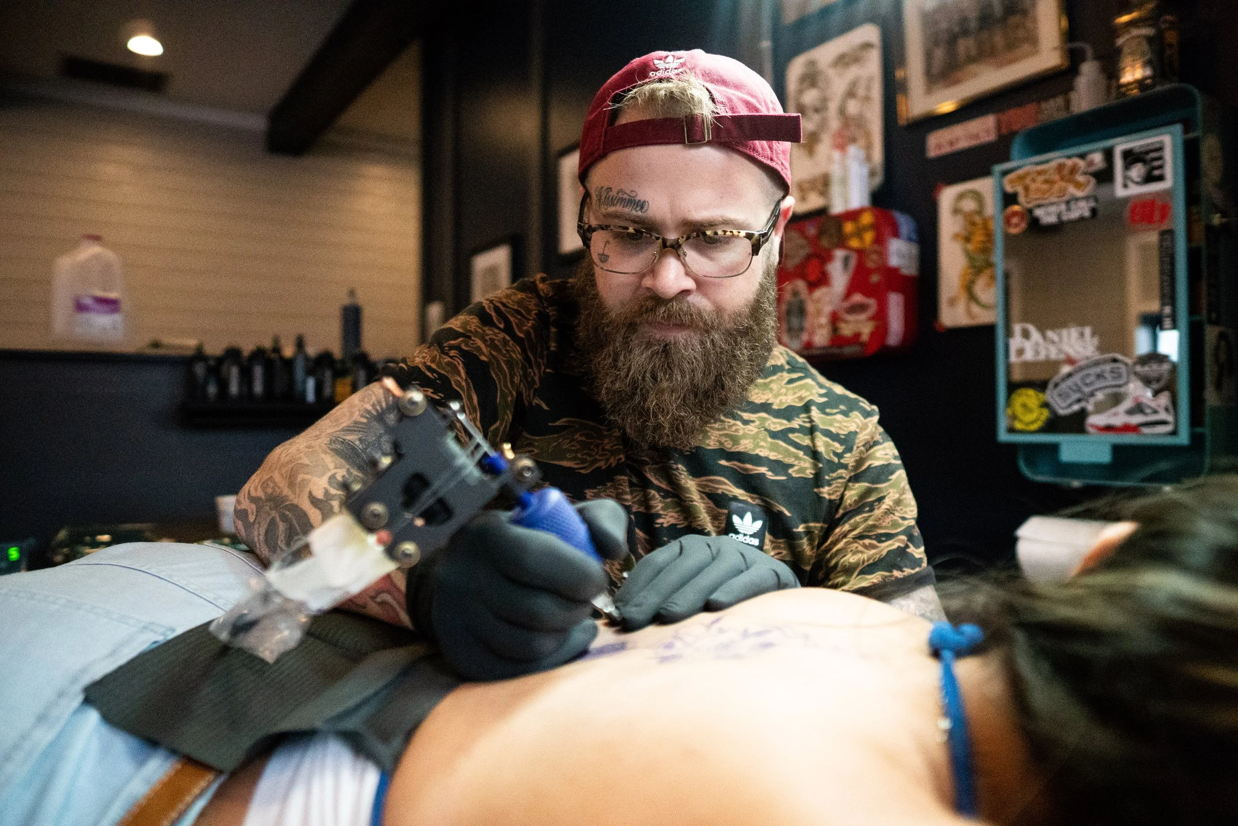 A tattoo artist wearing glasses, a red cap backwards, and gloves is working on a tattoo on a person's skin in a tattoo shop, with ink bottles and artwork visible in the background.