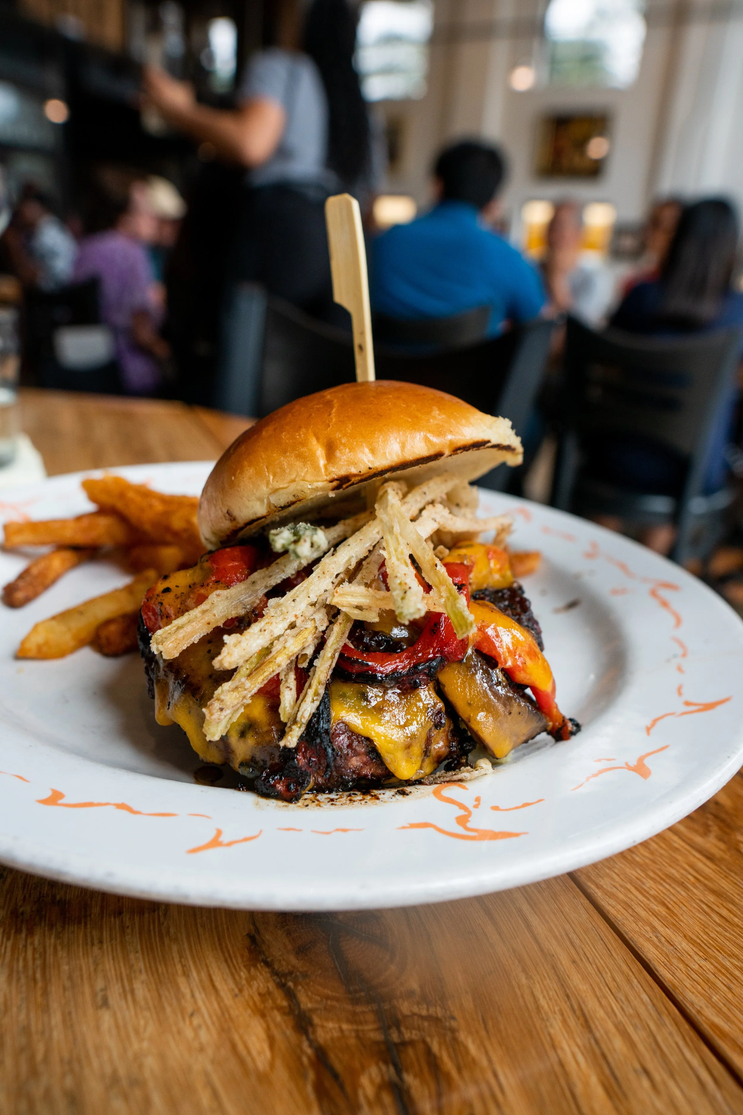 Close-up of a gourmet cheeseburger with fries on a white plate, with a restaurant scene in the background.