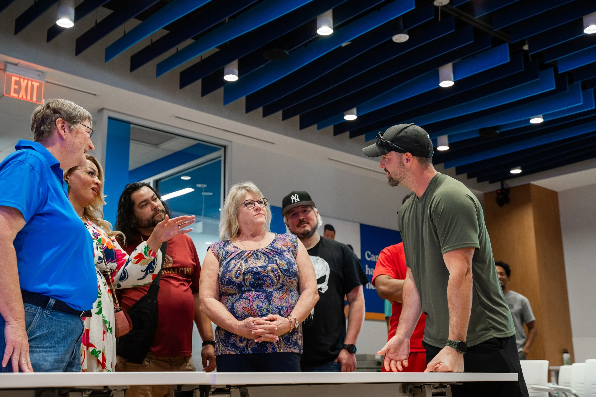 A group of six people standing around a table, engaged in a conversation, in an indoor setting with modern design elements, including blue ceiling beams and a glass wall.